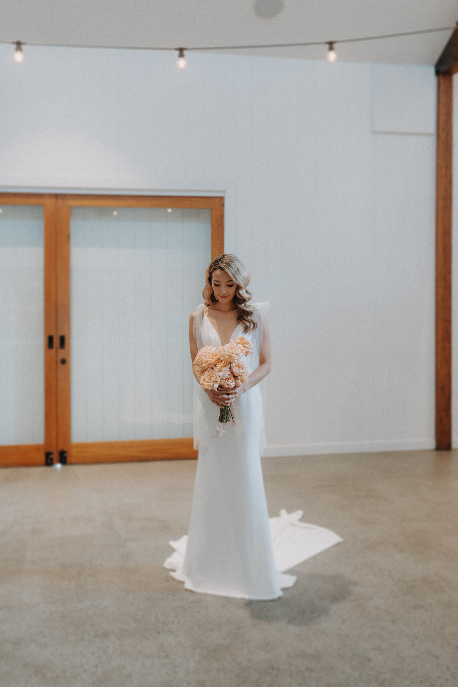 A bride in a white wedding dress holding a bouquet of peach roses stands indoors on a concrete floor with wooden-framed glass doors in the background.