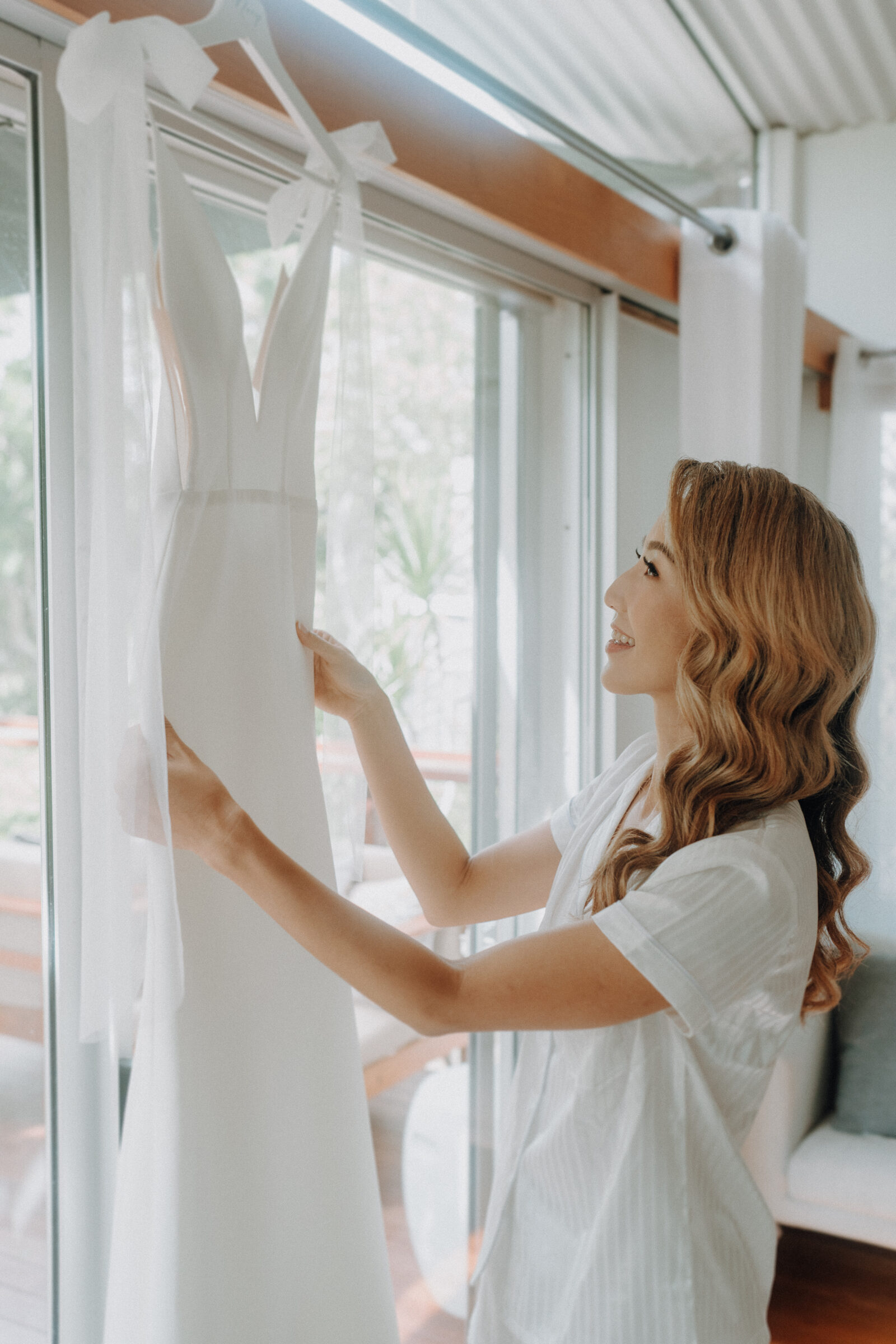 A woman with wavy hair smiles as she holds up a white wedding dress by a window with sheer curtains.