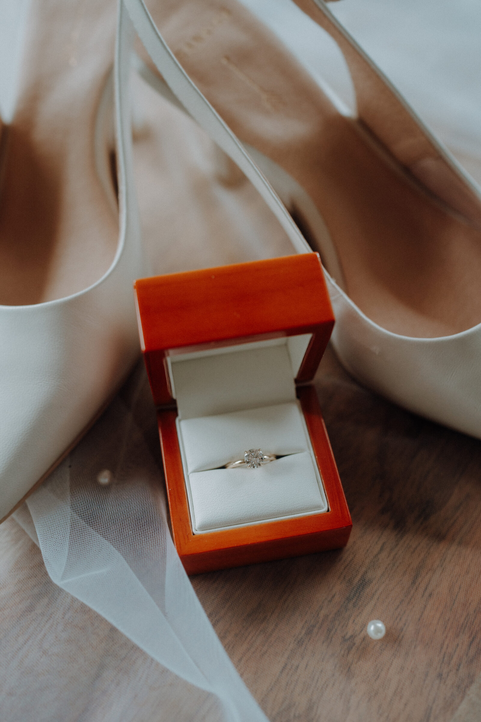 A diamond ring in a red box sits on a wooden surface, surrounded by white high heels and a sheer veil.