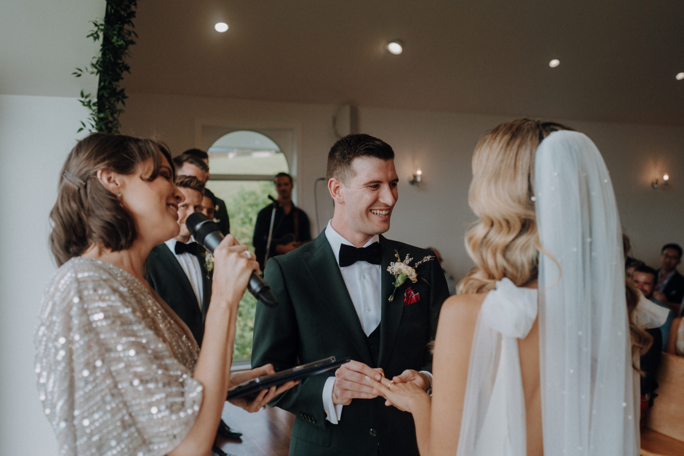 A bride and groom exchange rings during a wedding ceremony. The officiant, holding a microphone, stands beside them.