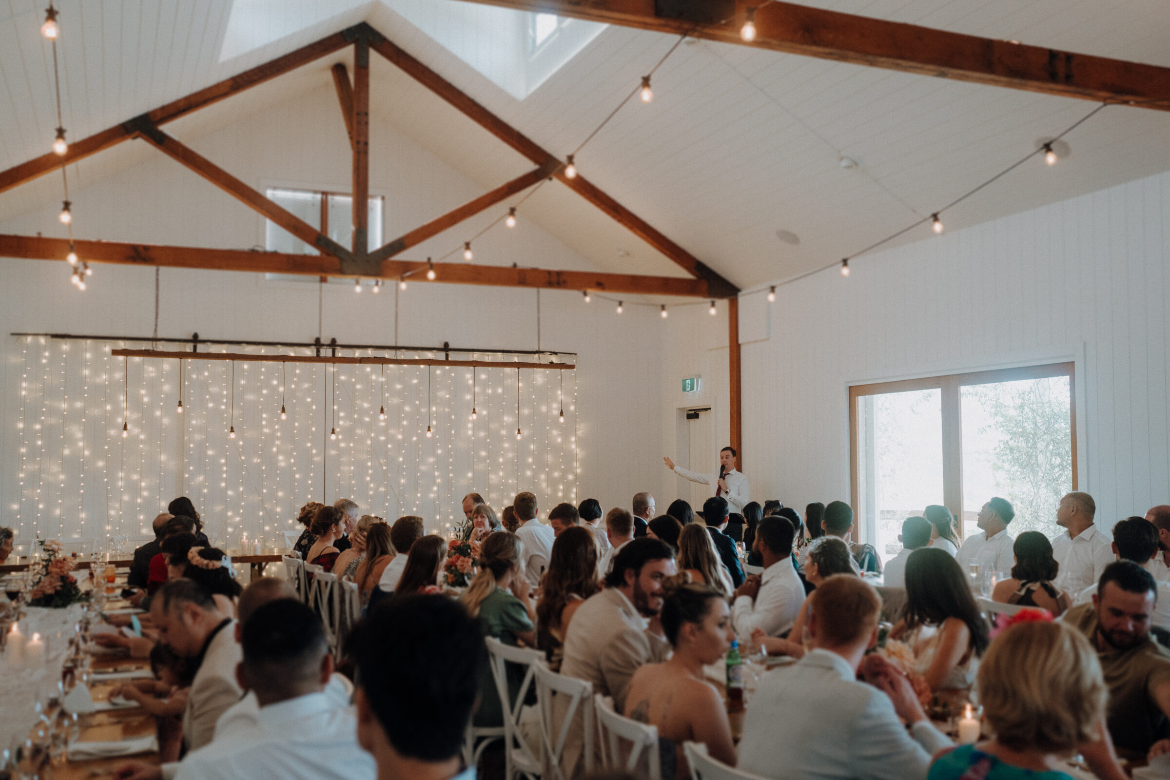 A person gives a speech to seated guests in a decorated hall with string lights and wooden beams.