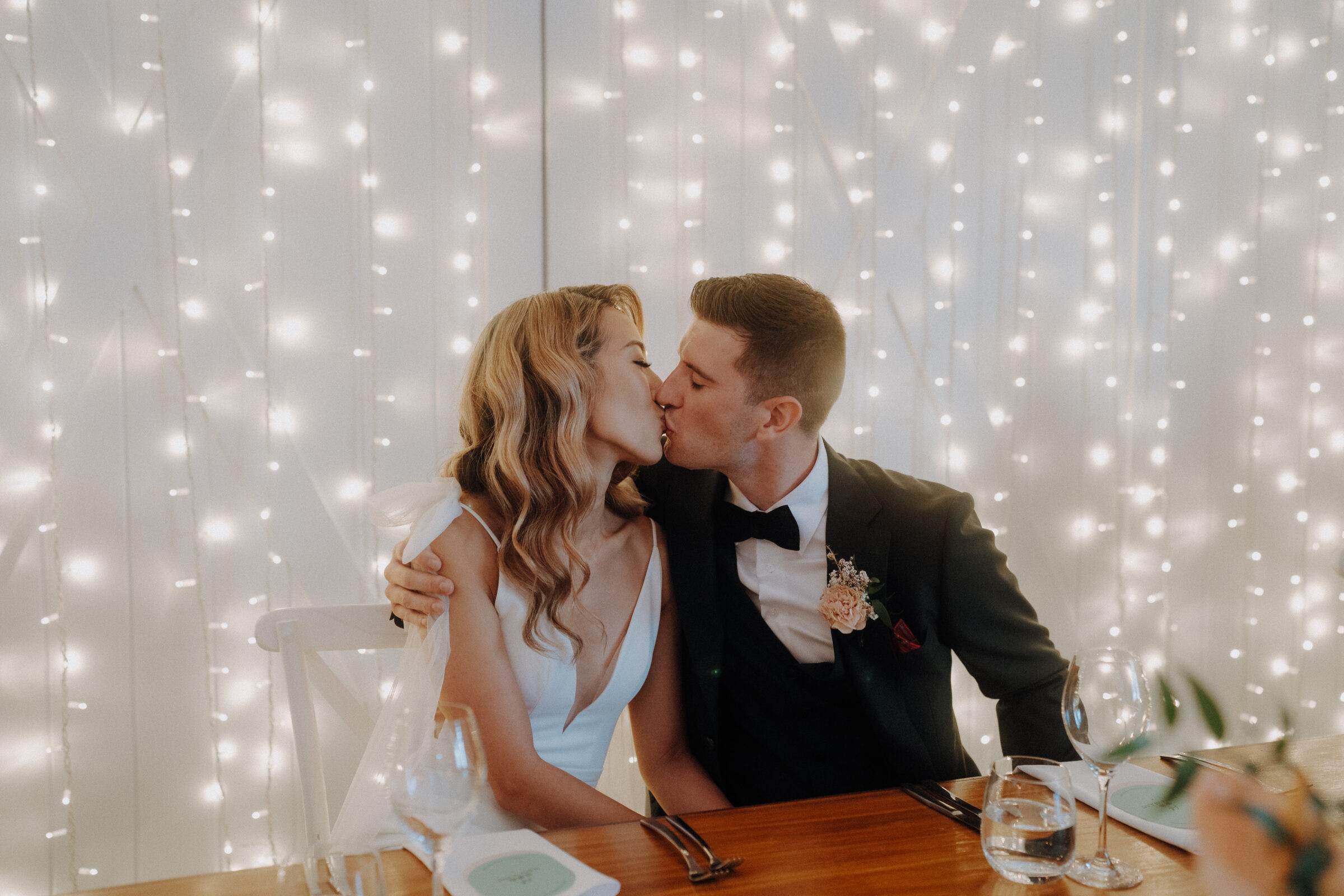 A couple in formal attire kisses while seated at a table, with a backdrop of fairy lights.