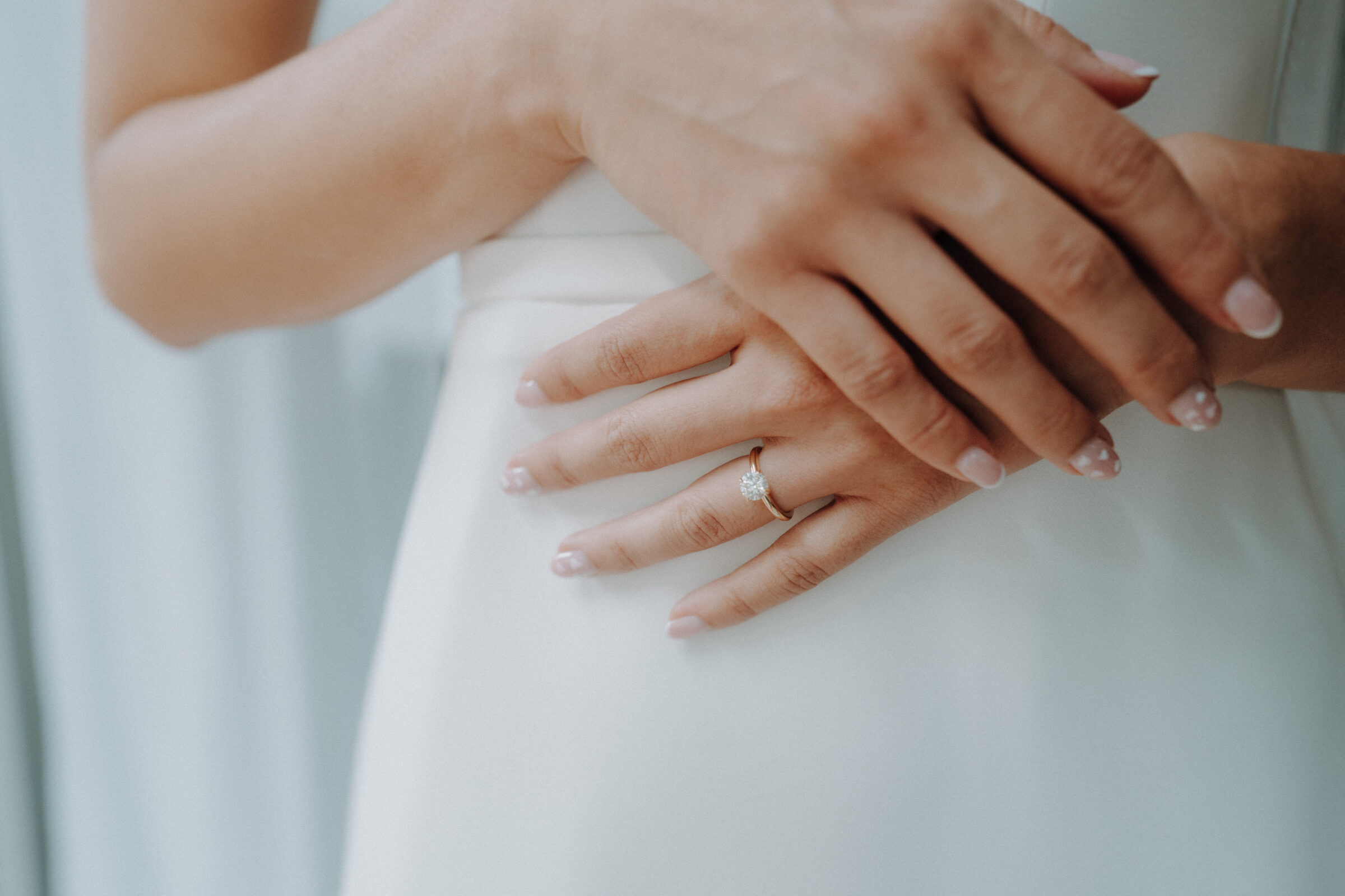 Close-up of a person wearing a white dress with hands clasped, showing a diamond ring on the left hand.