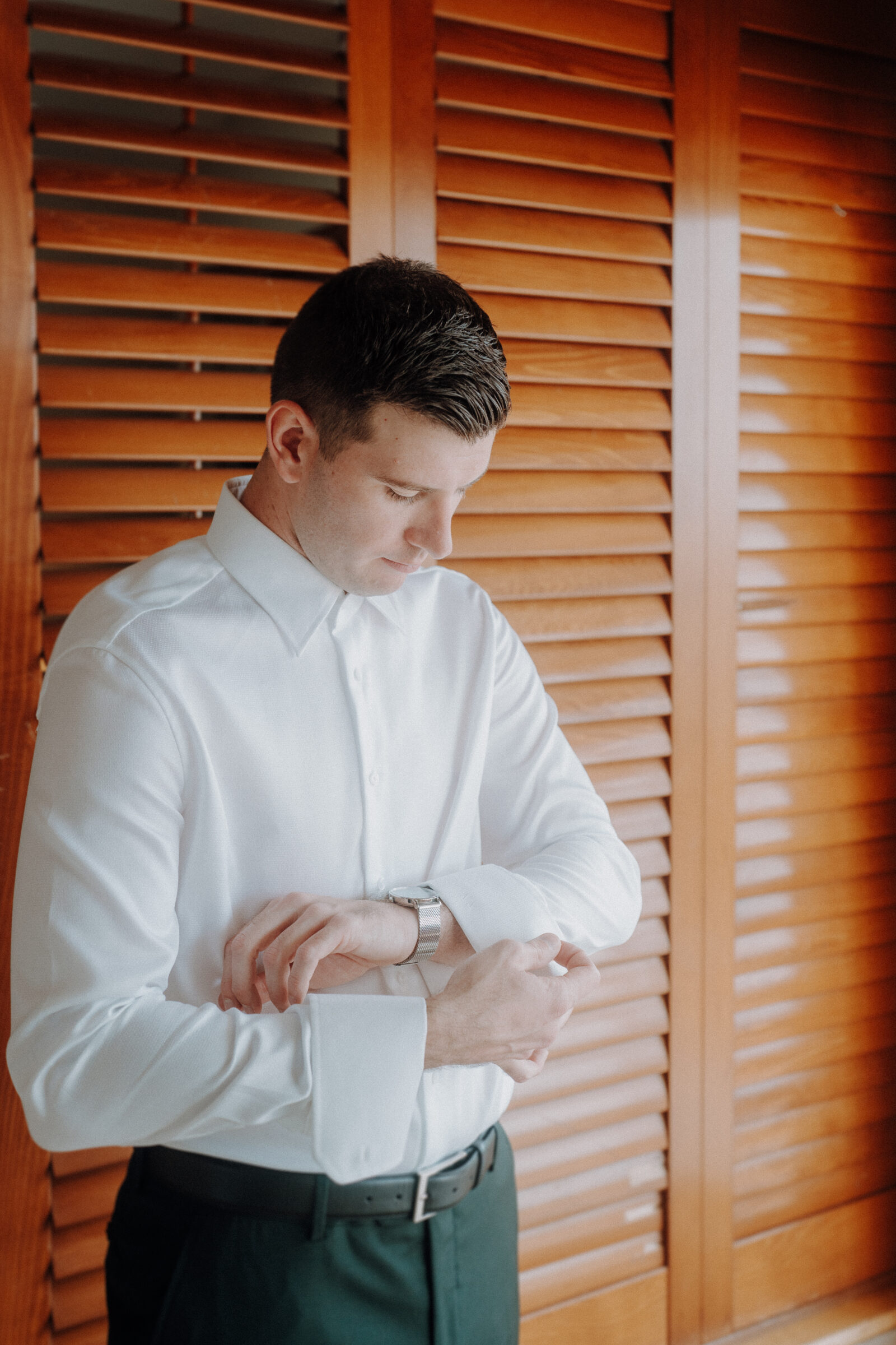 Man in a white shirt adjusts his watch, standing in front of wooden shutters.