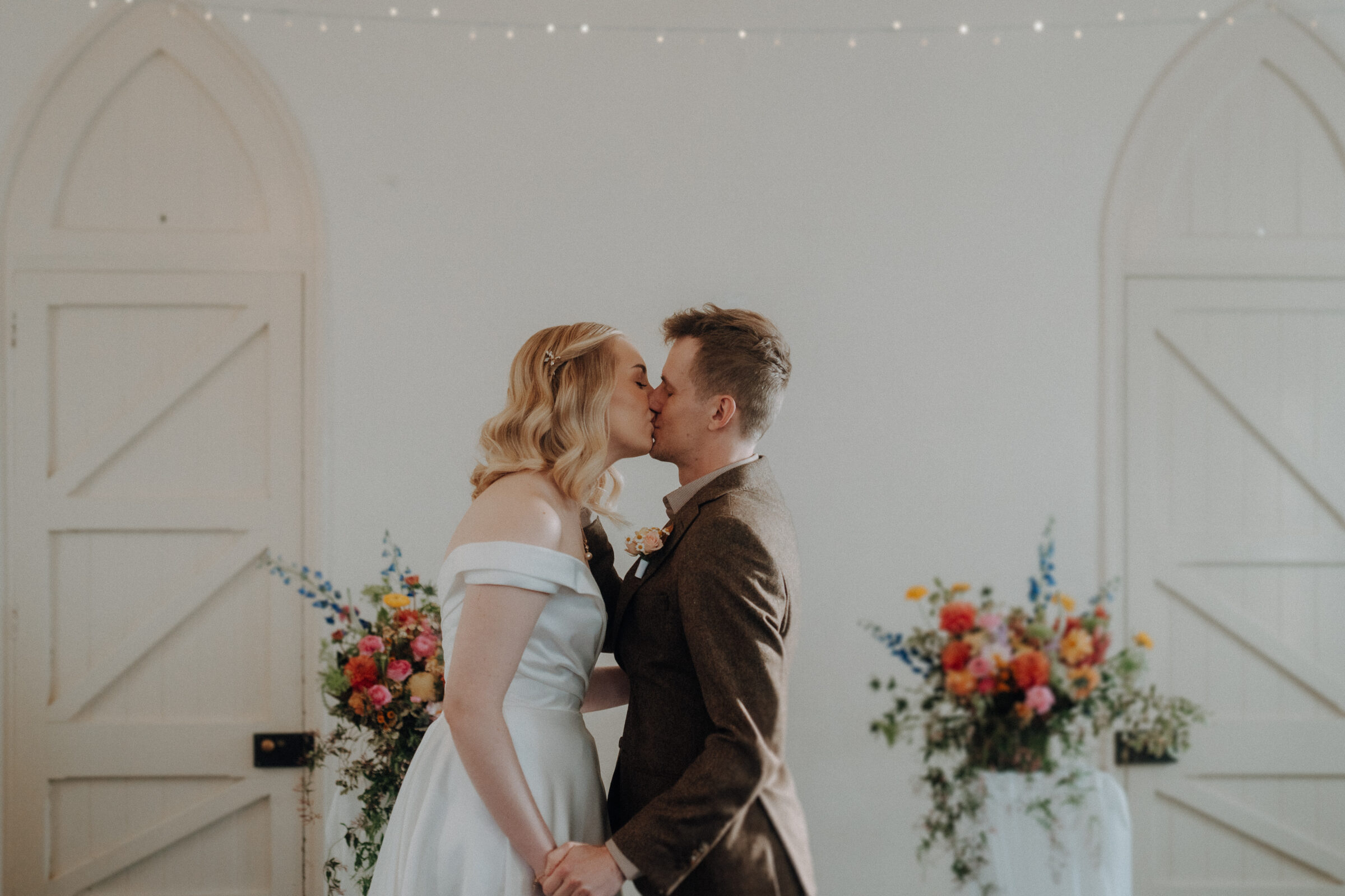 A couple kisses at a wedding ceremony with floral arrangements and arched doors in the background.