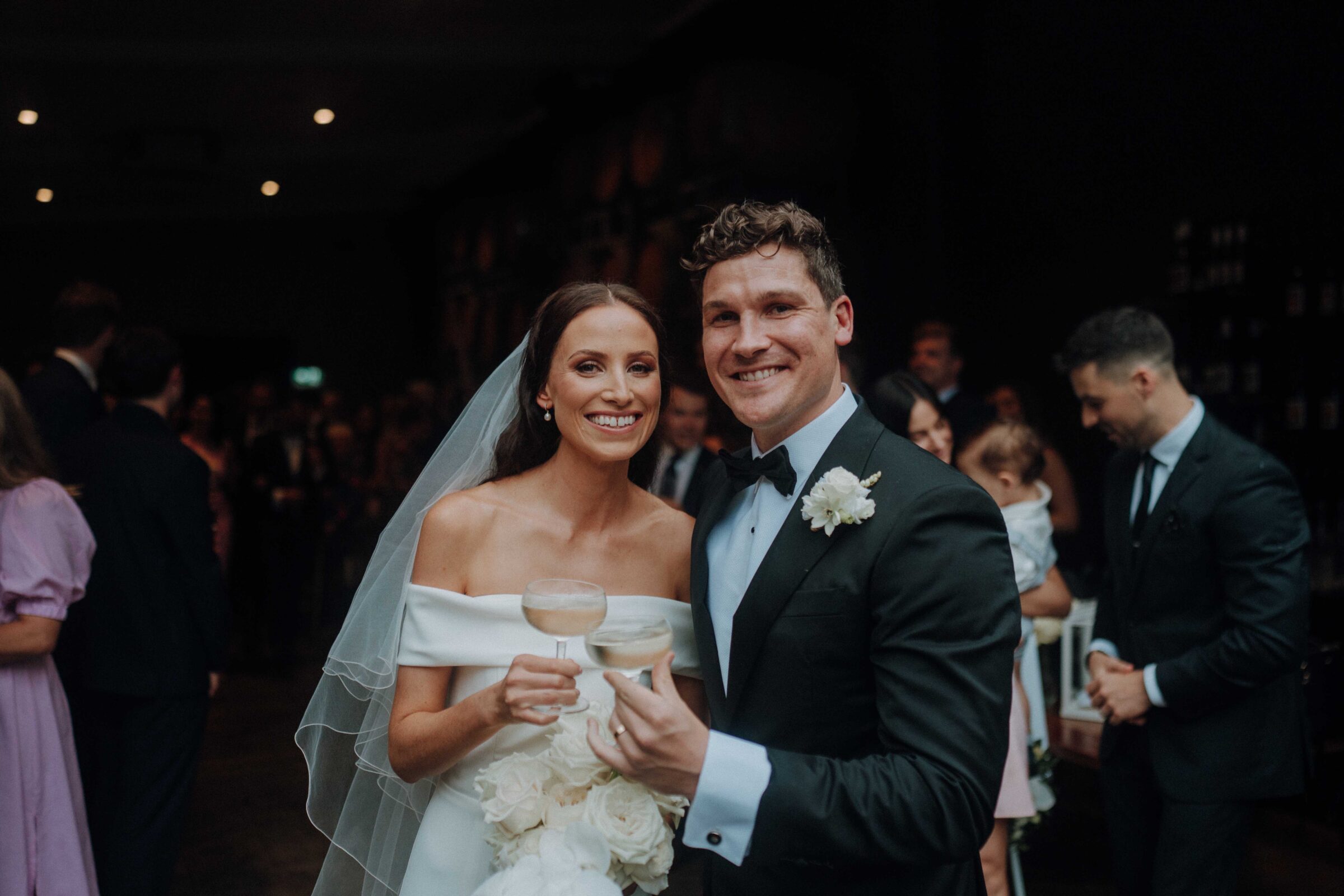 Bride and groom smiling and holding drinks at a wedding reception, with guests in the background.