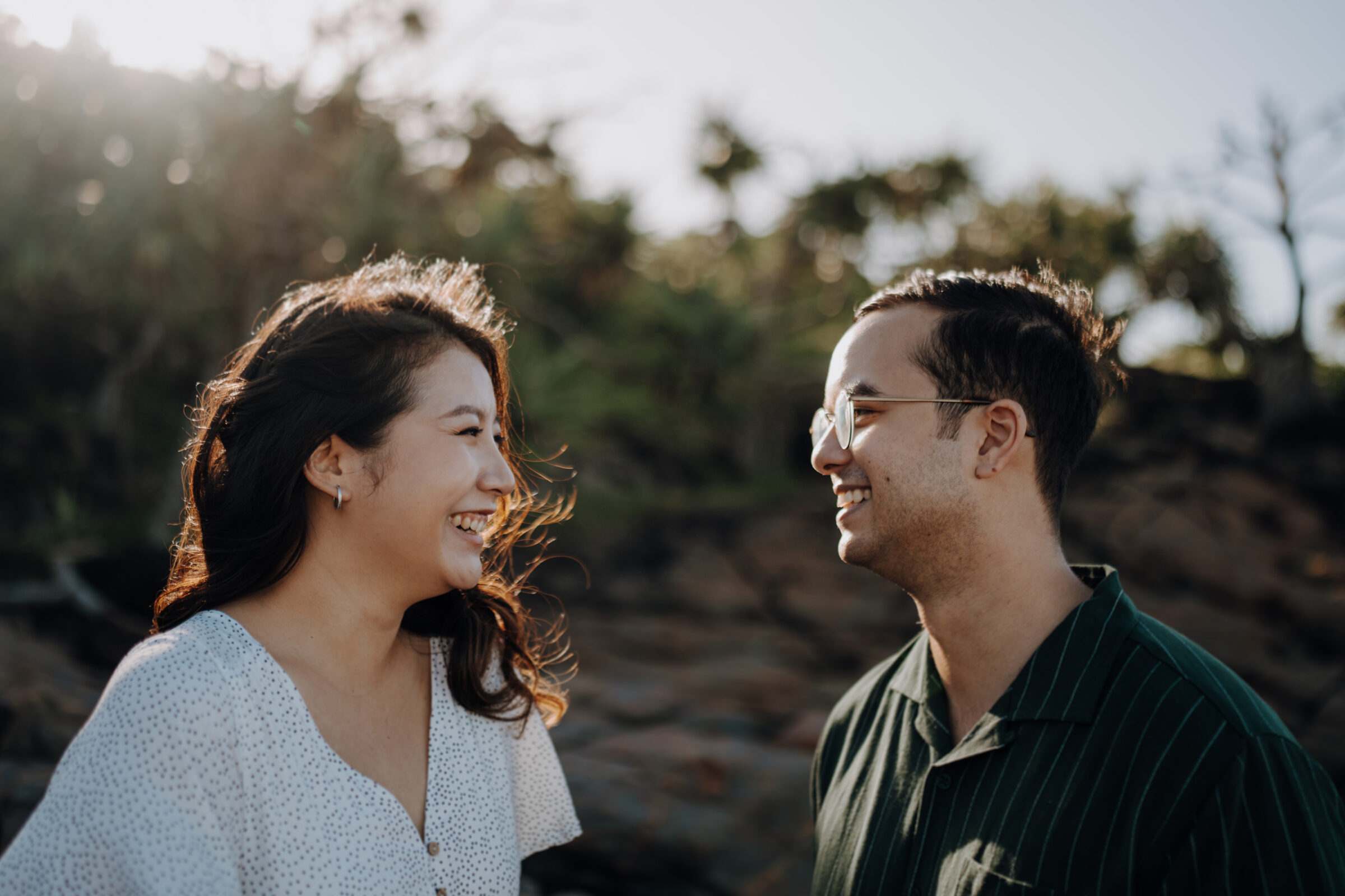 A man and a woman stand outdoors, smiling at each other. The background features greenery and rocks under a clear sky.