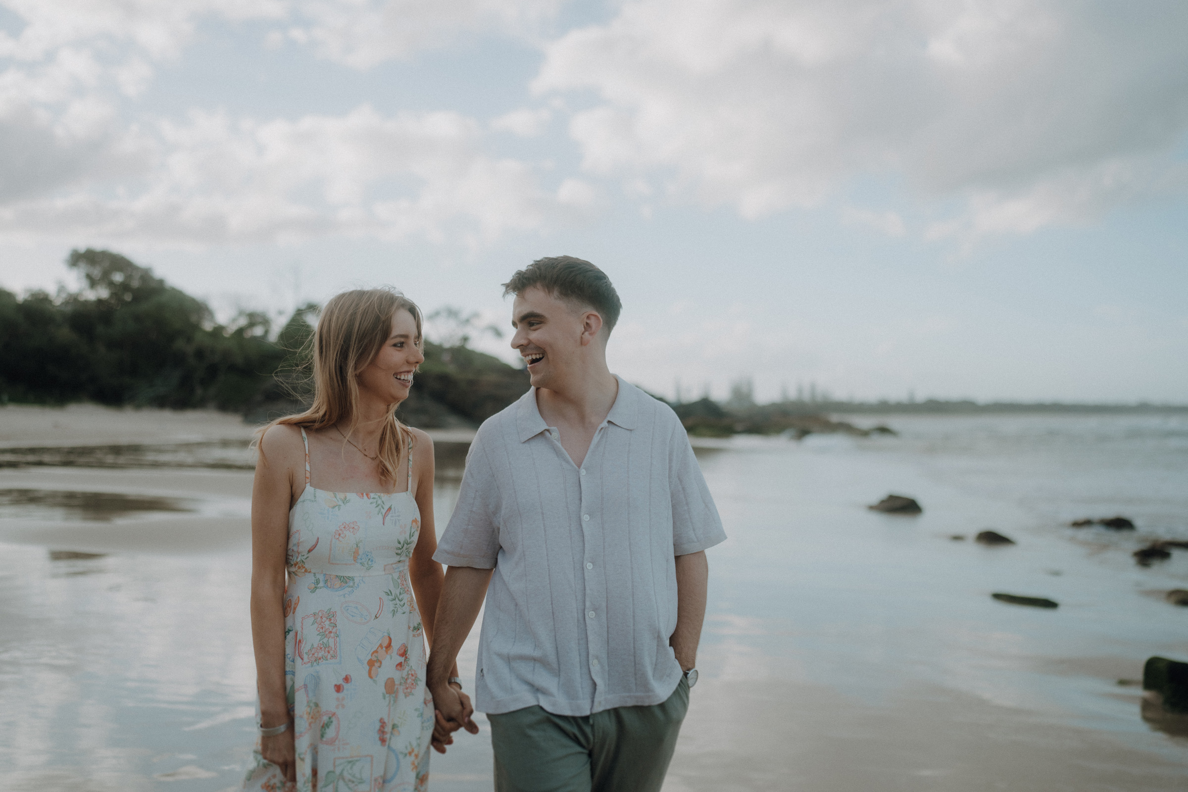 A couple holds hands, smiling at each other, while walking on a beach with rocks and trees in the background under a cloudy sky.
