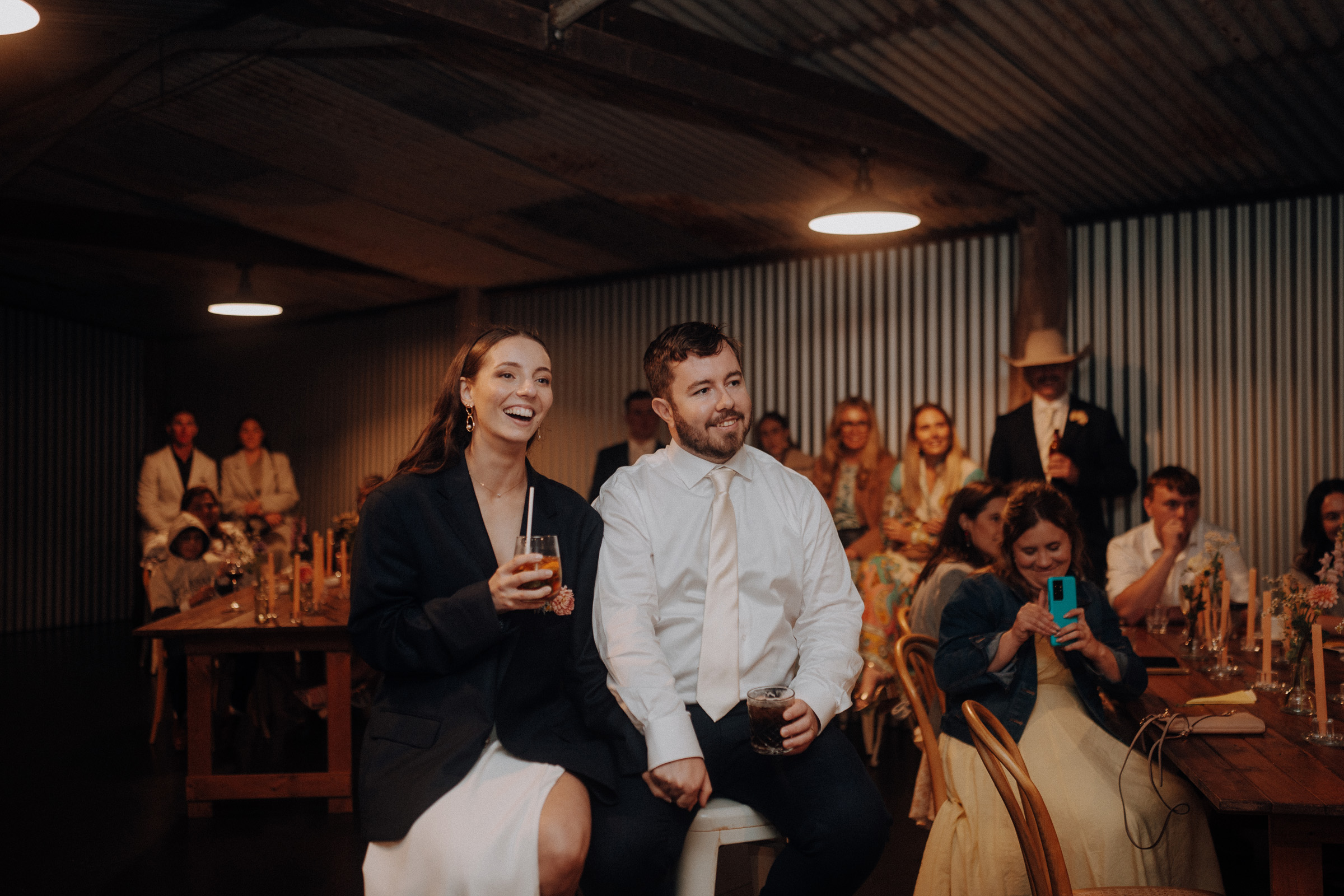 A couple sits together at an indoor event, smiling and holding drinks. People are seated and standing in the background, watching an unseen event.