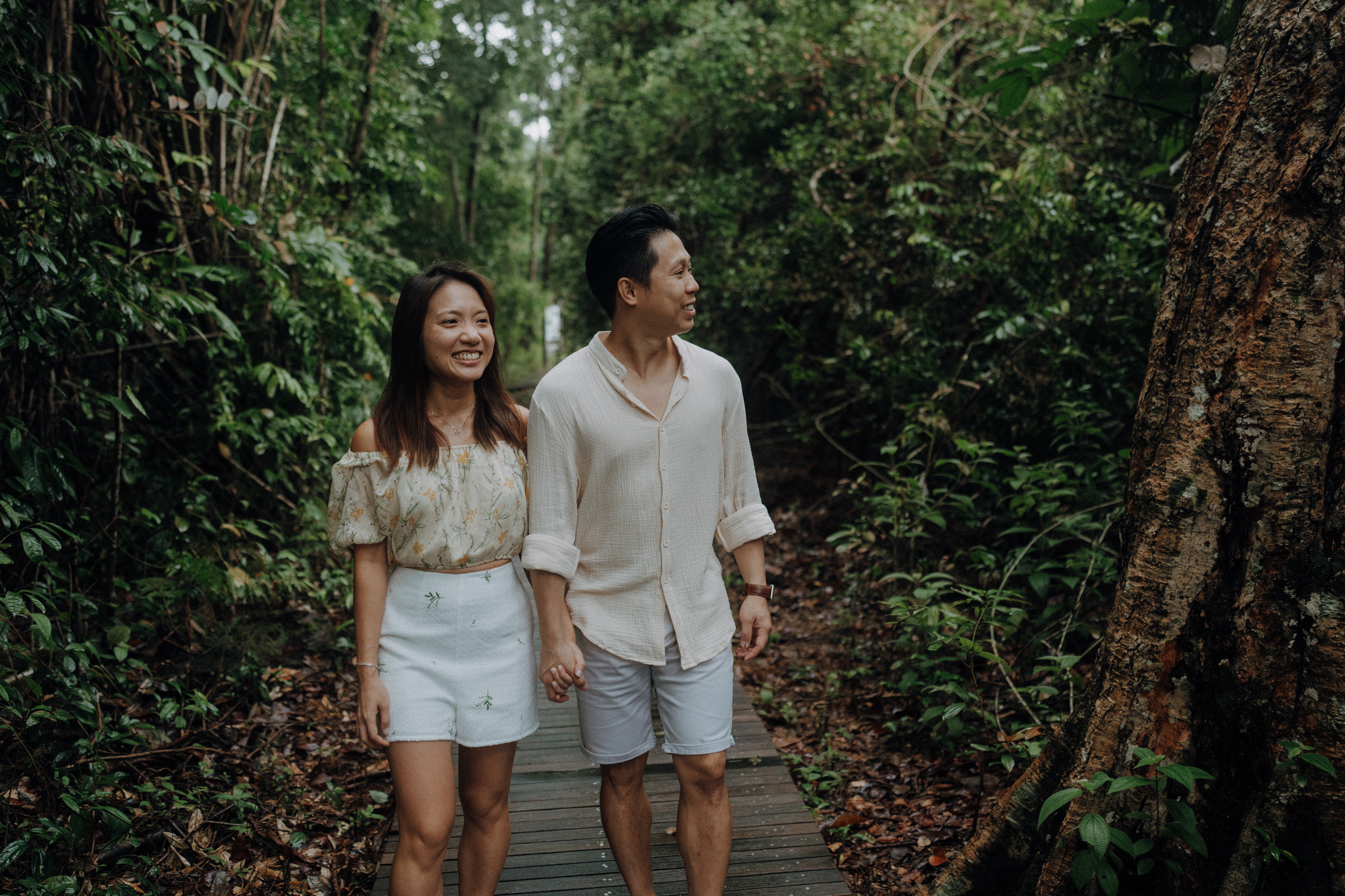 A couple walks hand in hand on a wooden path through a dense, green forest. They are wearing casual summer clothes and appear to be enjoying the nature around them.