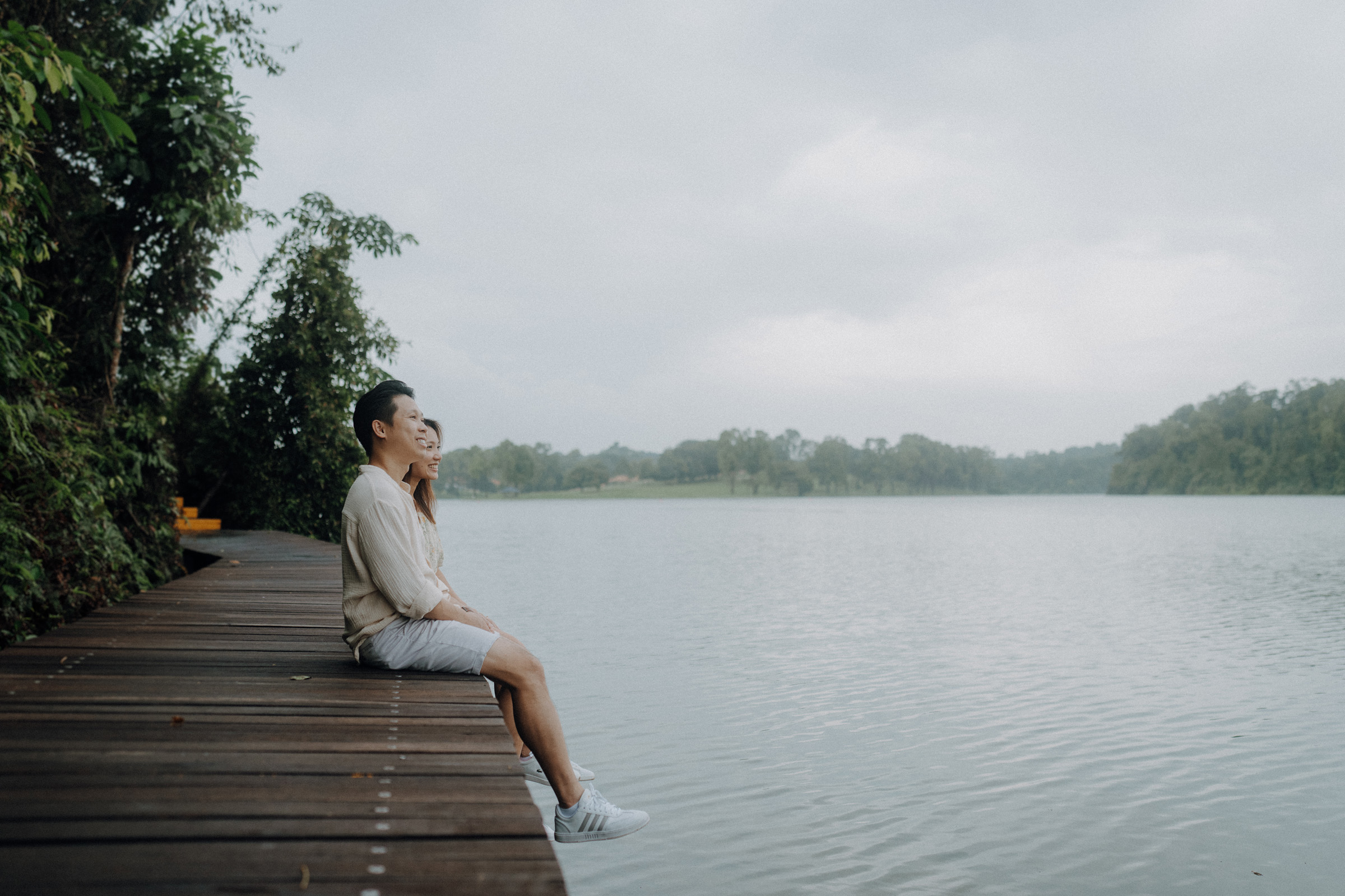 A couple sits on a wooden dock beside a calm lake, surrounded by greenery, under an overcast sky.