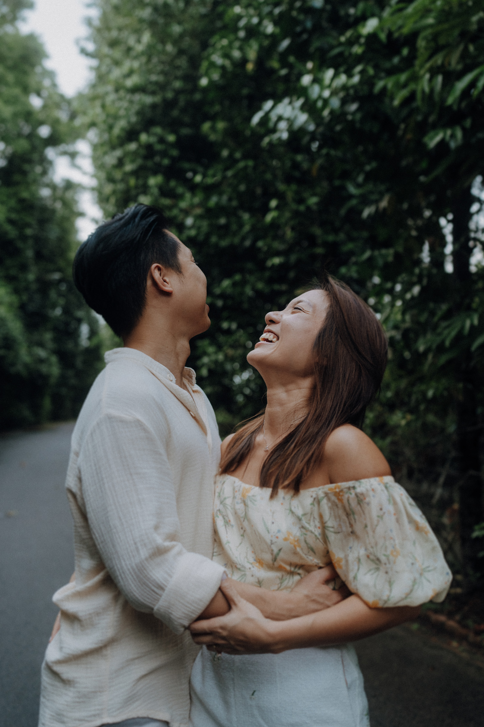 A couple laughs together on a tree-lined path. Both wear light-colored clothing, and the setting is lush and green.