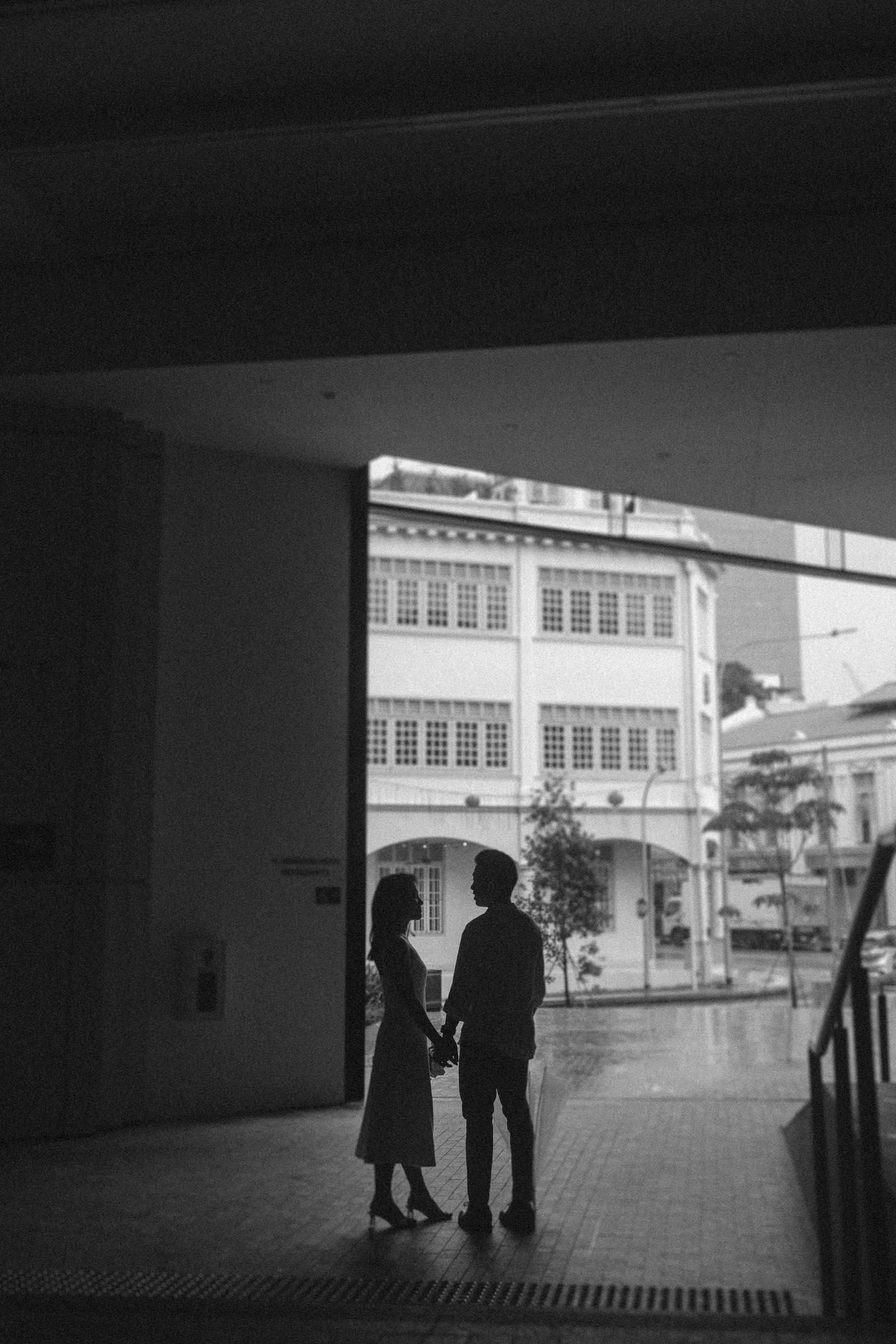 Silhouetted couple holding hands, standing in a building's entrance with street and buildings visible outside on a rainy day.