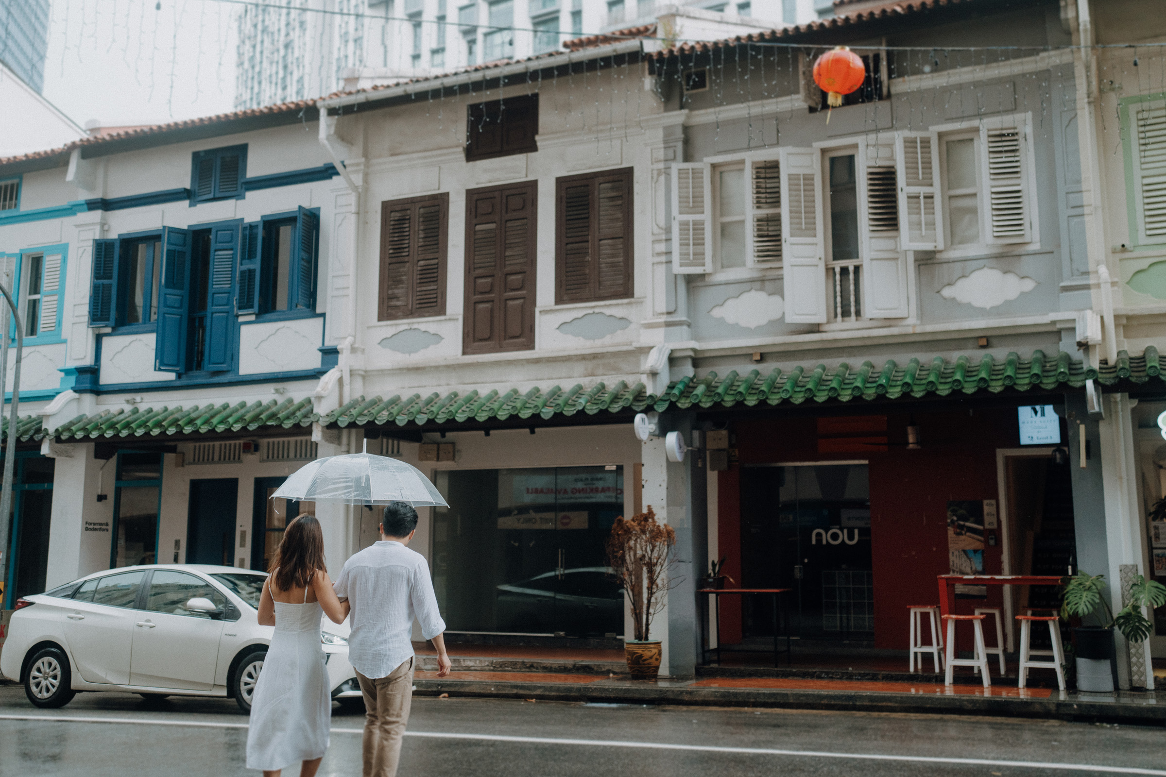 A couple walks with an umbrella down a rainy street lined with colorful, traditional shophouses. A white car is parked on the road.