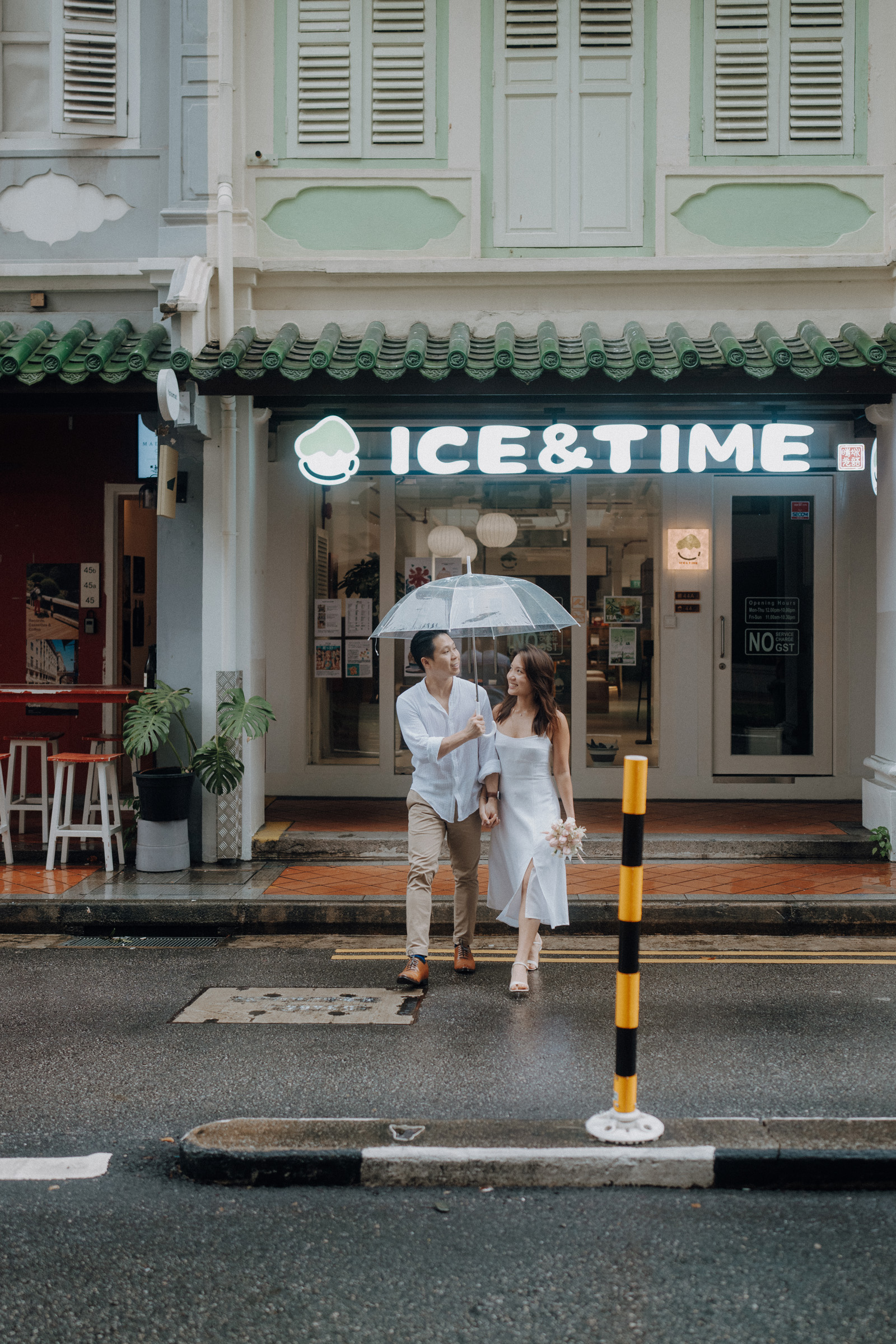 A couple walks under an umbrella on a wet street in front of an "ICE & TIME" storefront.