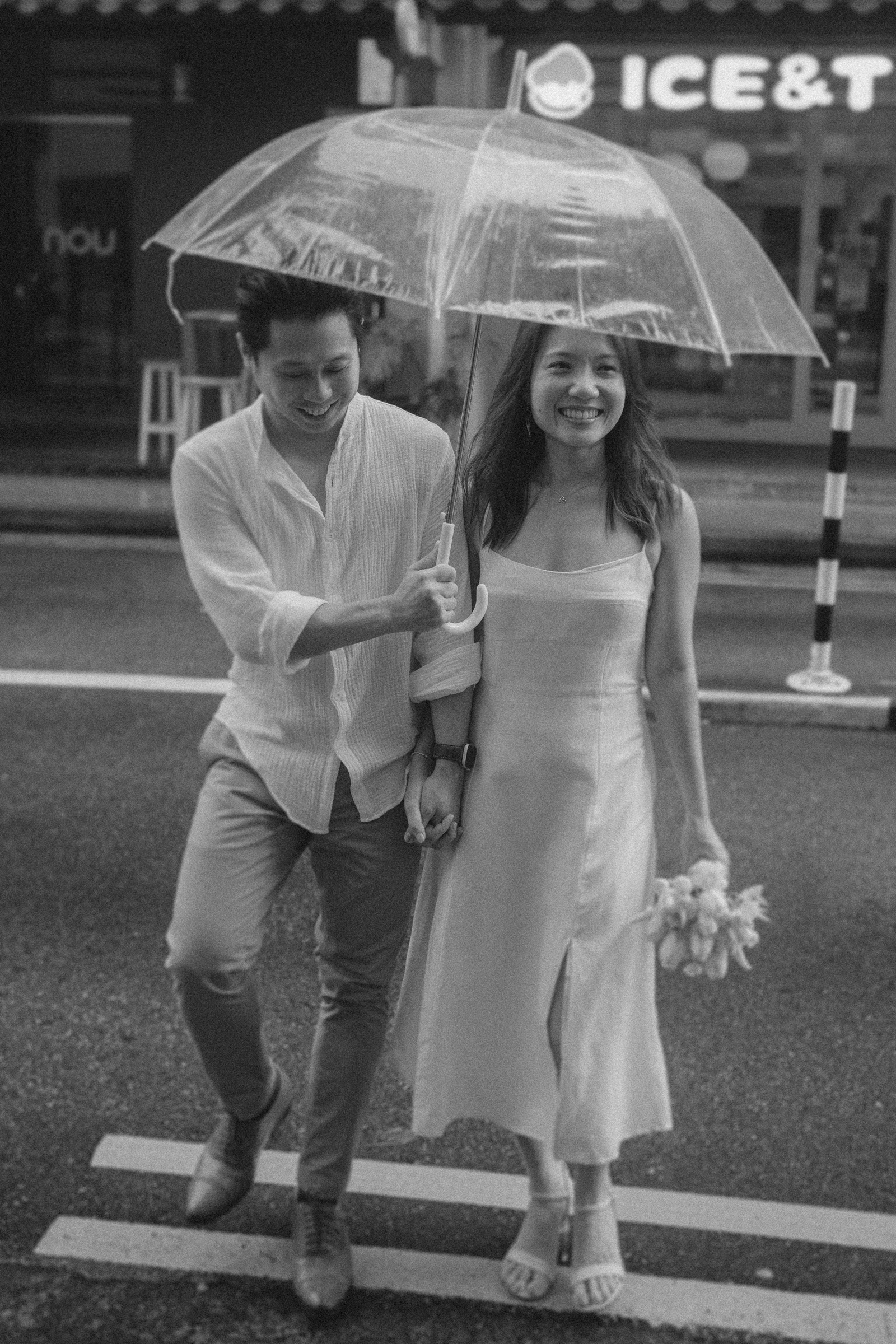 A couple walks hand in hand under a clear umbrella on a wet street, with the woman holding flowers and both smiling.