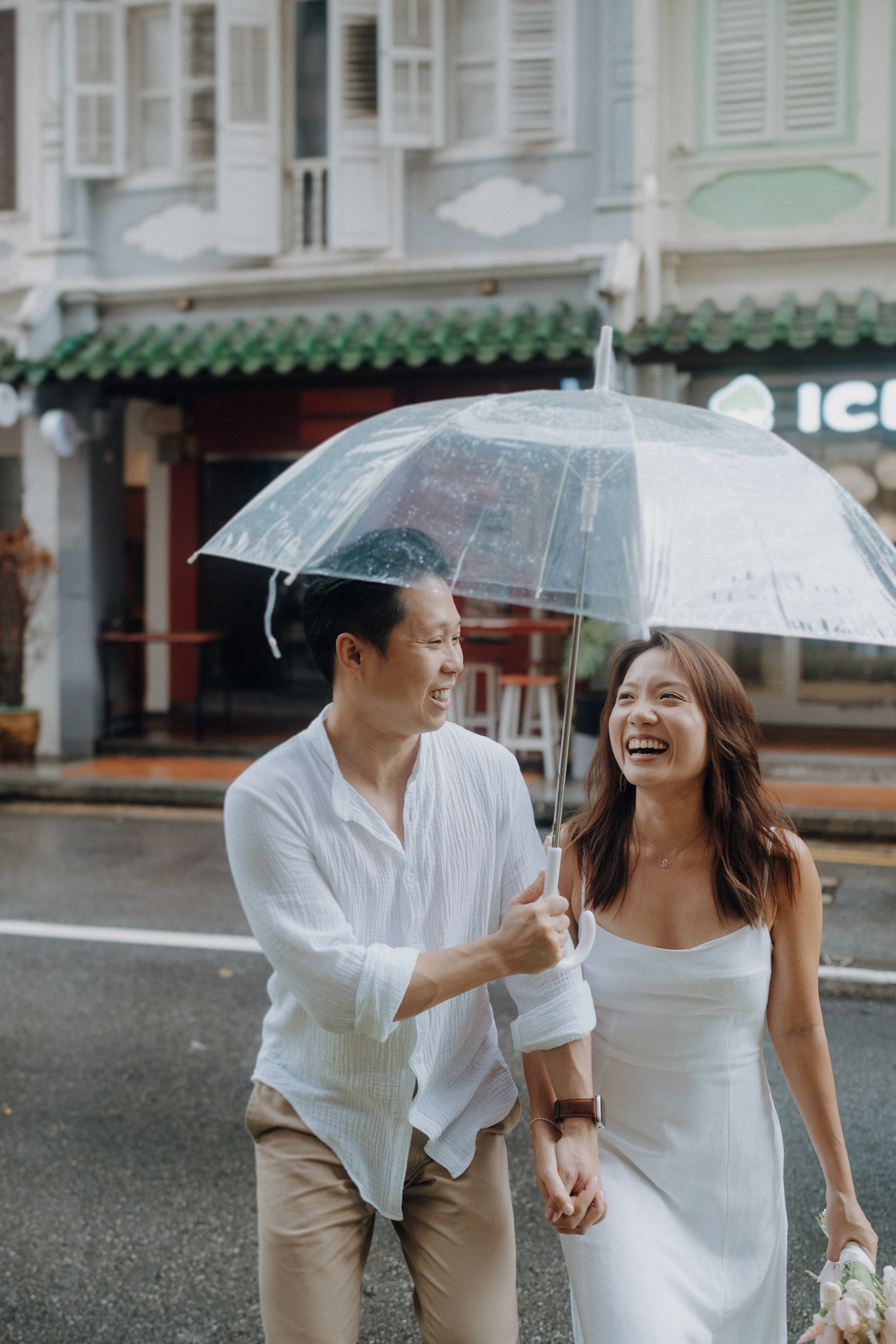 A couple walks happily under a clear umbrella on a rainy street, with pastel-colored buildings in the background.