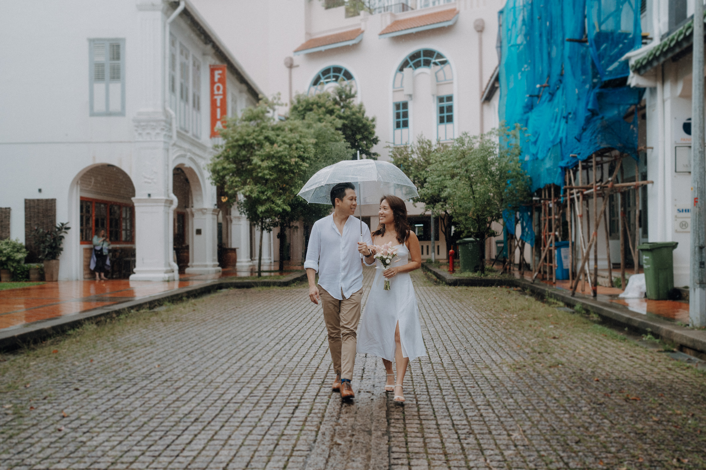 A couple walks together under an umbrella on a cobblestone street, surrounded by buildings and greenery on a rainy day.