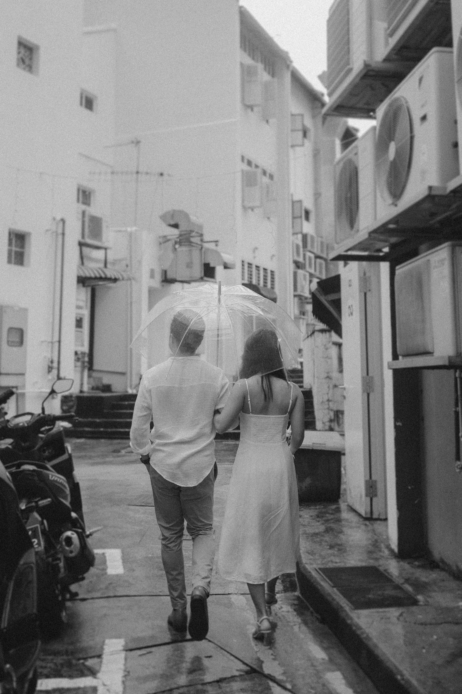 A couple walks under a transparent umbrella in a narrow alley, surrounded by buildings and parked scooters. The scene is in black and white.