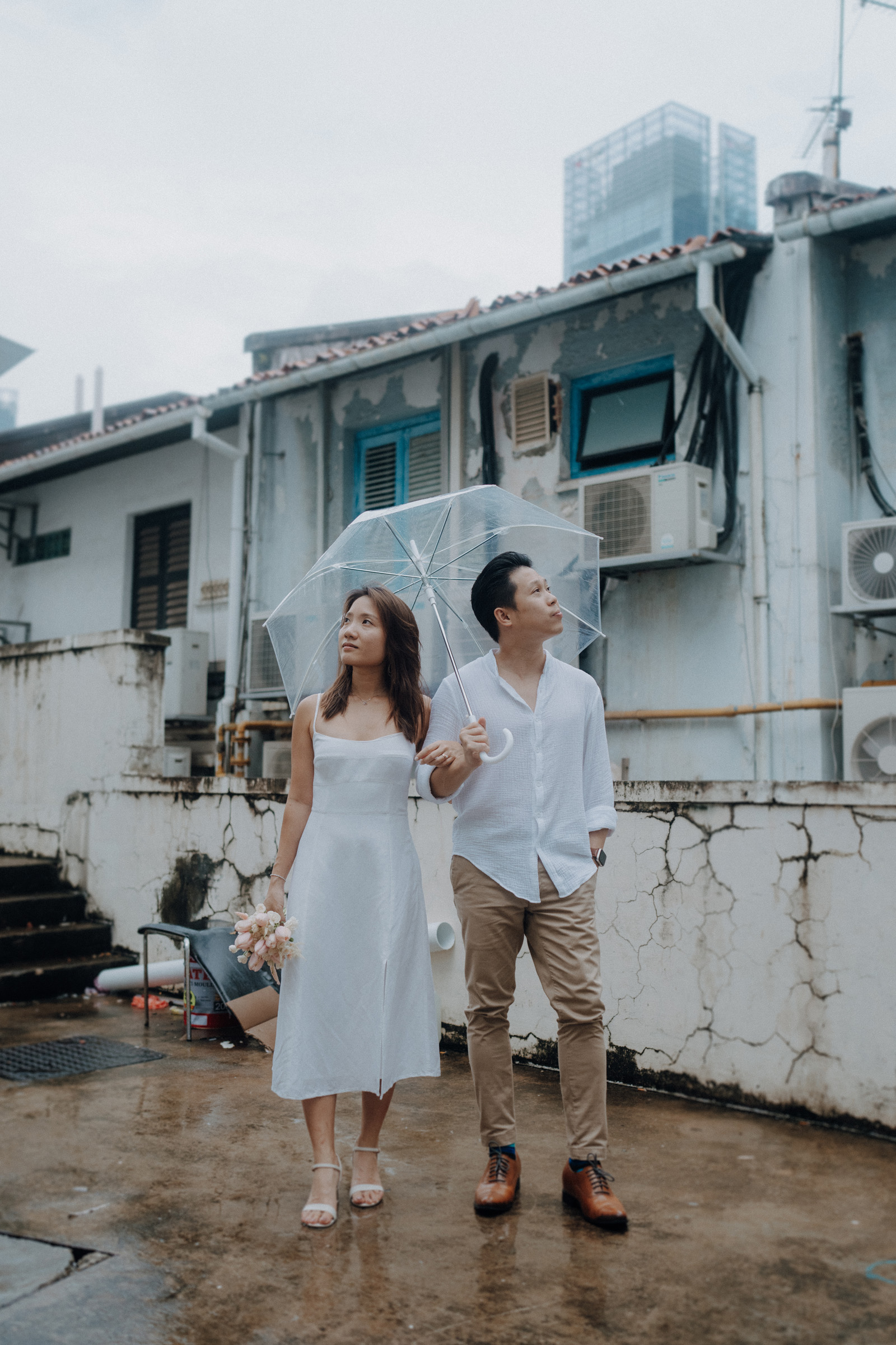 A couple stands under a clear umbrella in front of an aged building, looking upward. The woman holds a bouquet, and both wear casual, light-colored clothing.