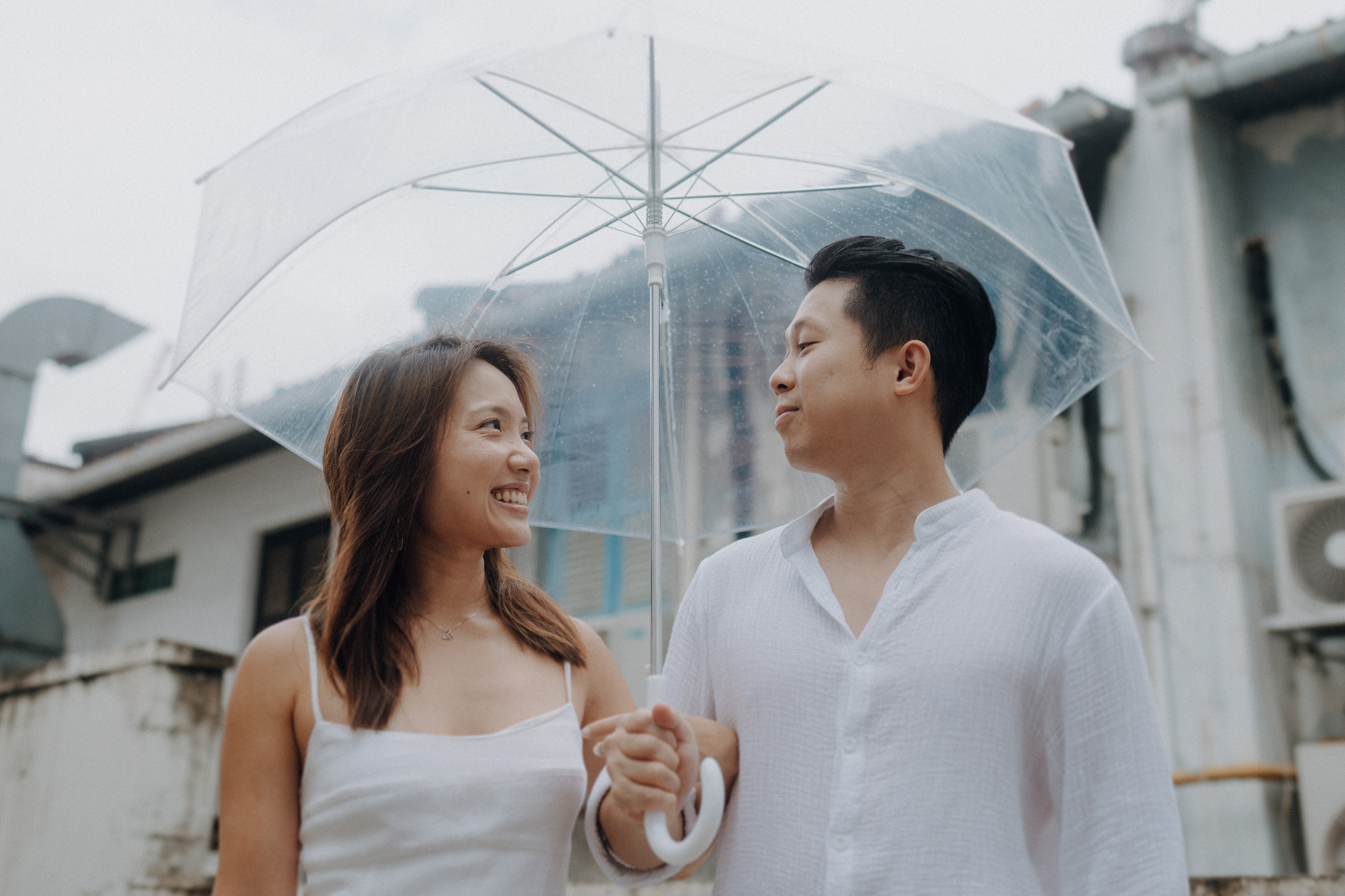 A couple in white clothing smiles at each other under a clear umbrella on a cloudy day.