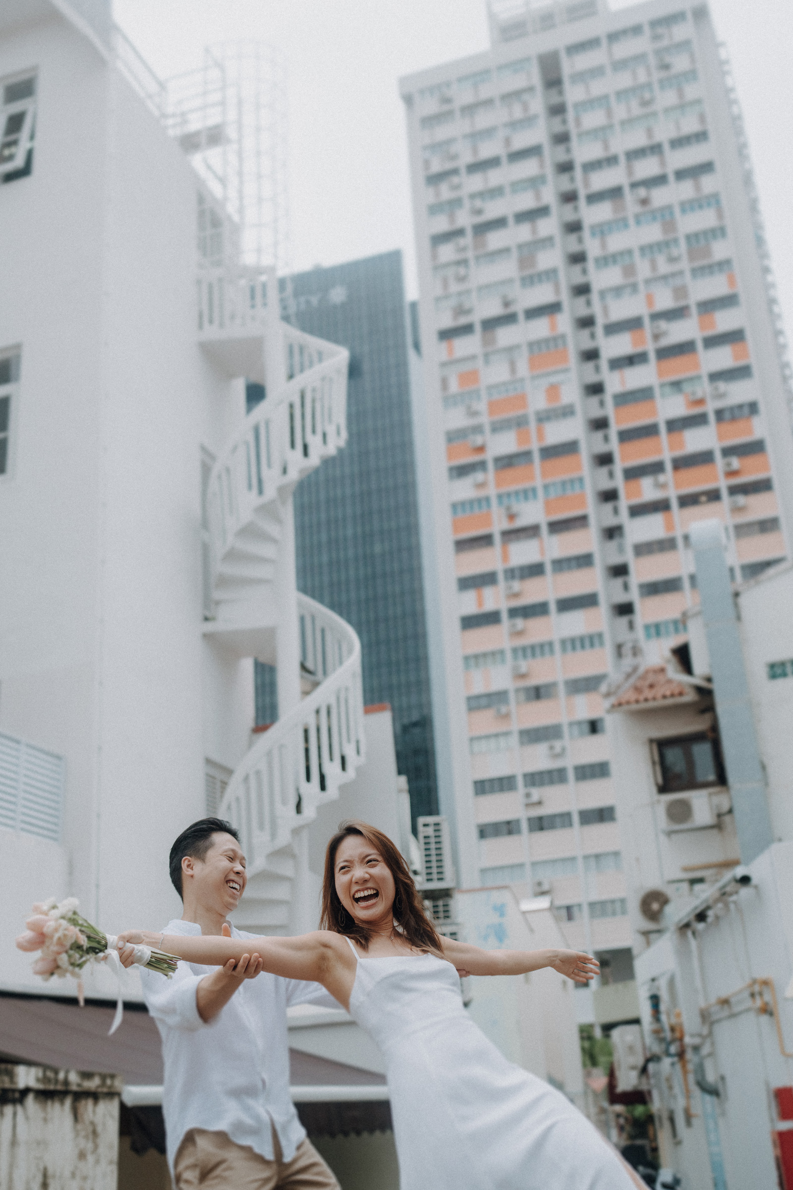 A couple joyfully holding hands, the woman extending her arms, with a spiral staircase and tall buildings in the background.