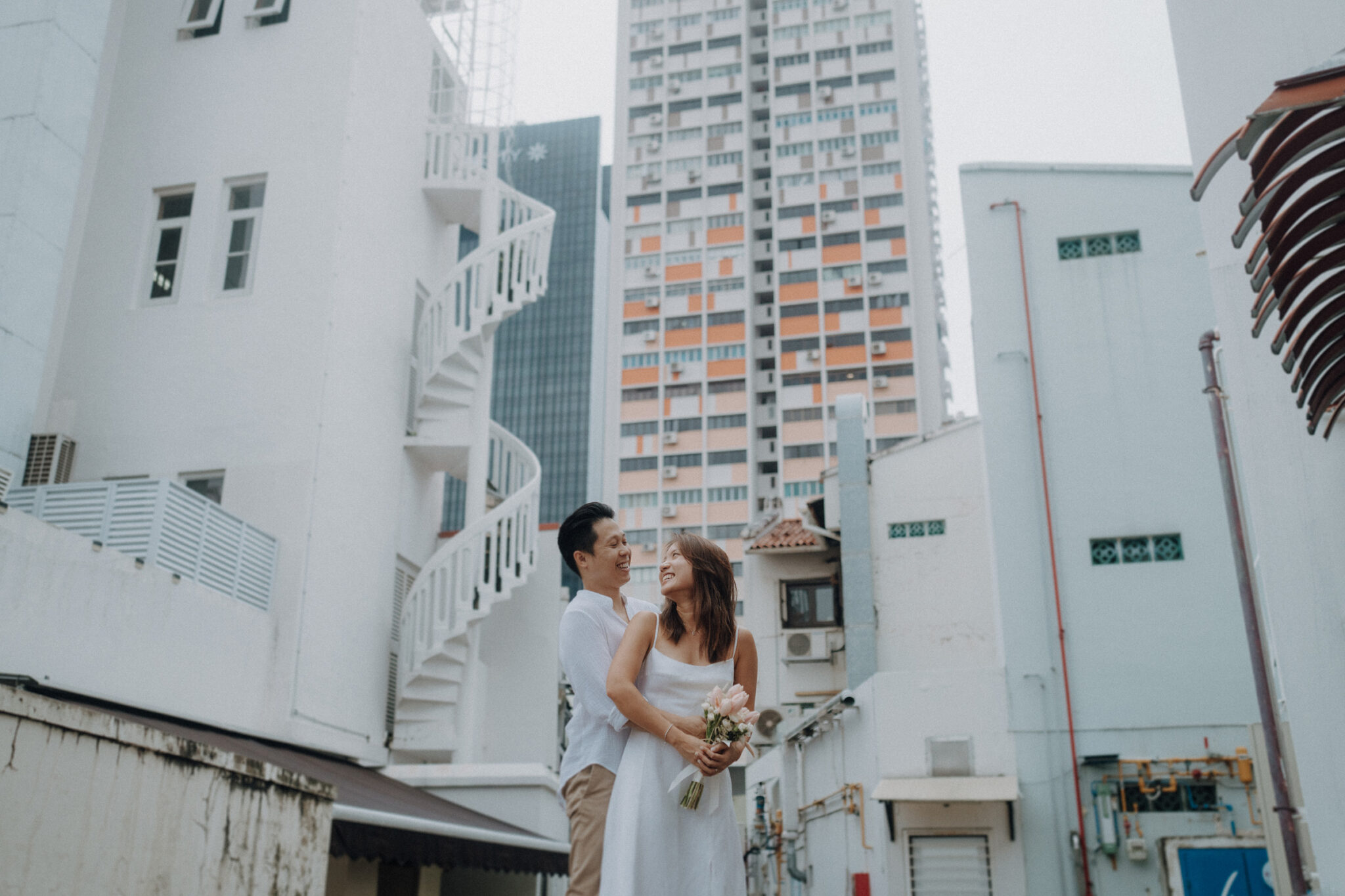 A couple stands in an urban alley, with tall buildings in the background. The woman holds a bouquet of flowers.