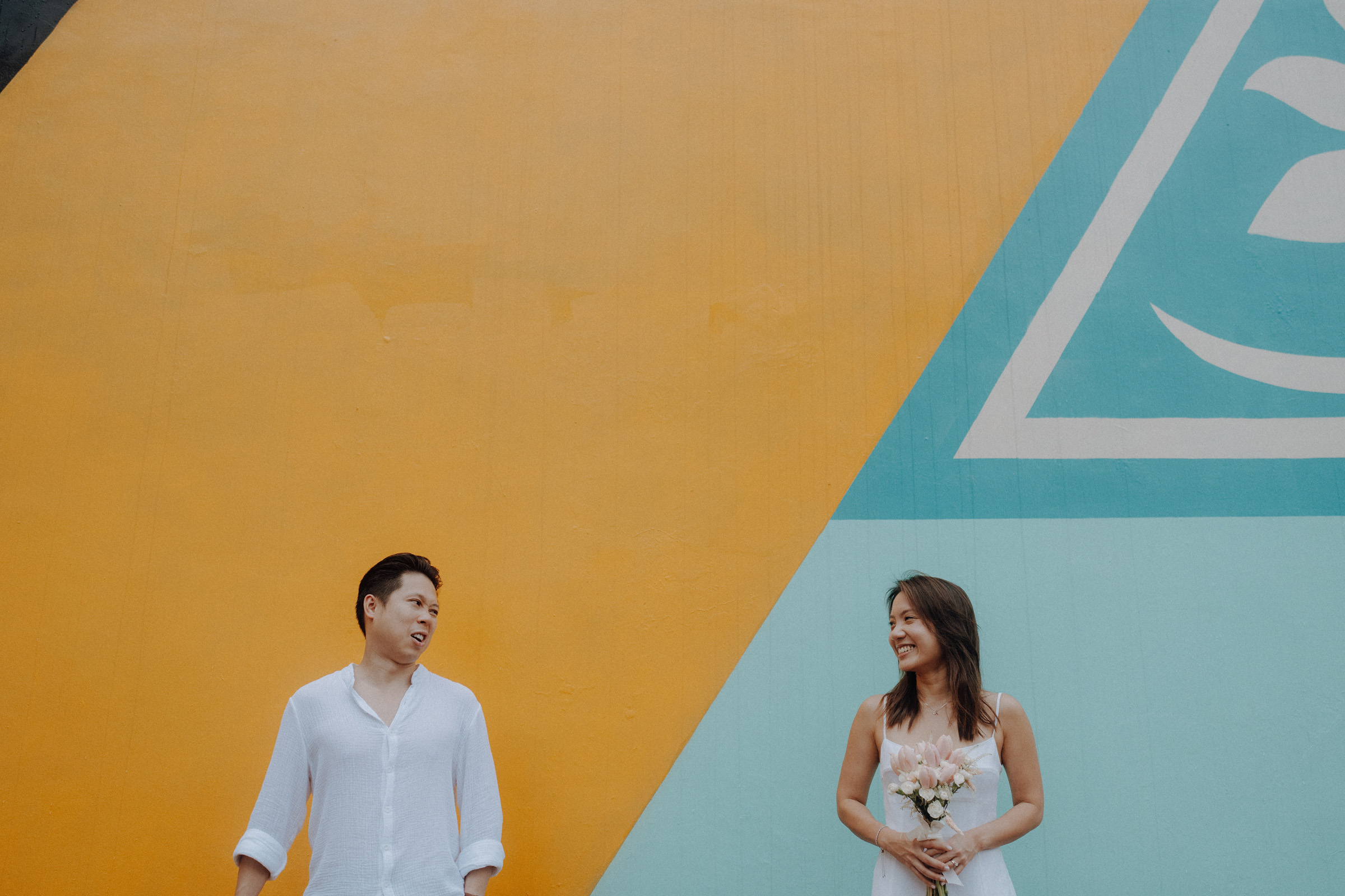 A man and woman stand apart in front of a geometric mural. The woman holds flowers and they both smile.