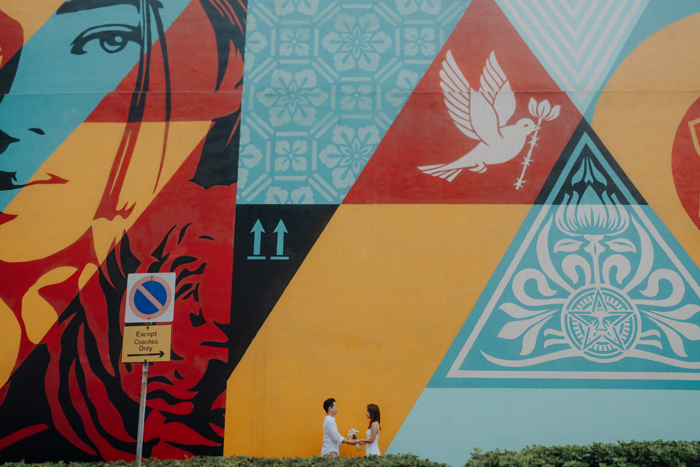 Two people stand side by side in front of a colorful mural with geometric shapes, a bird, and a woman's face. A sign reads "Except Cycles Only.