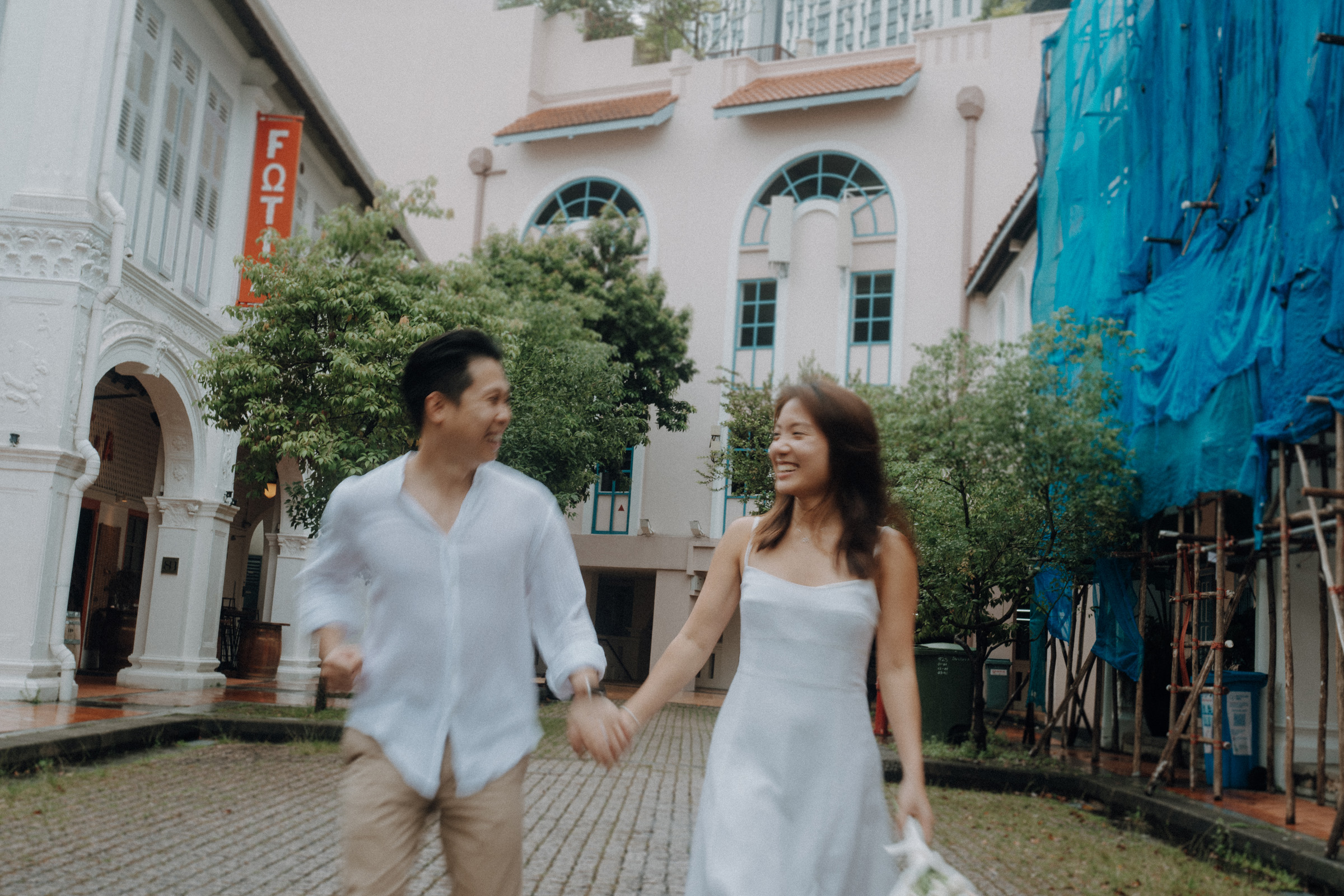 A couple holds hands, smiling and walking on a cobblestone path between two buildings, with trees and construction scaffolding in the background.