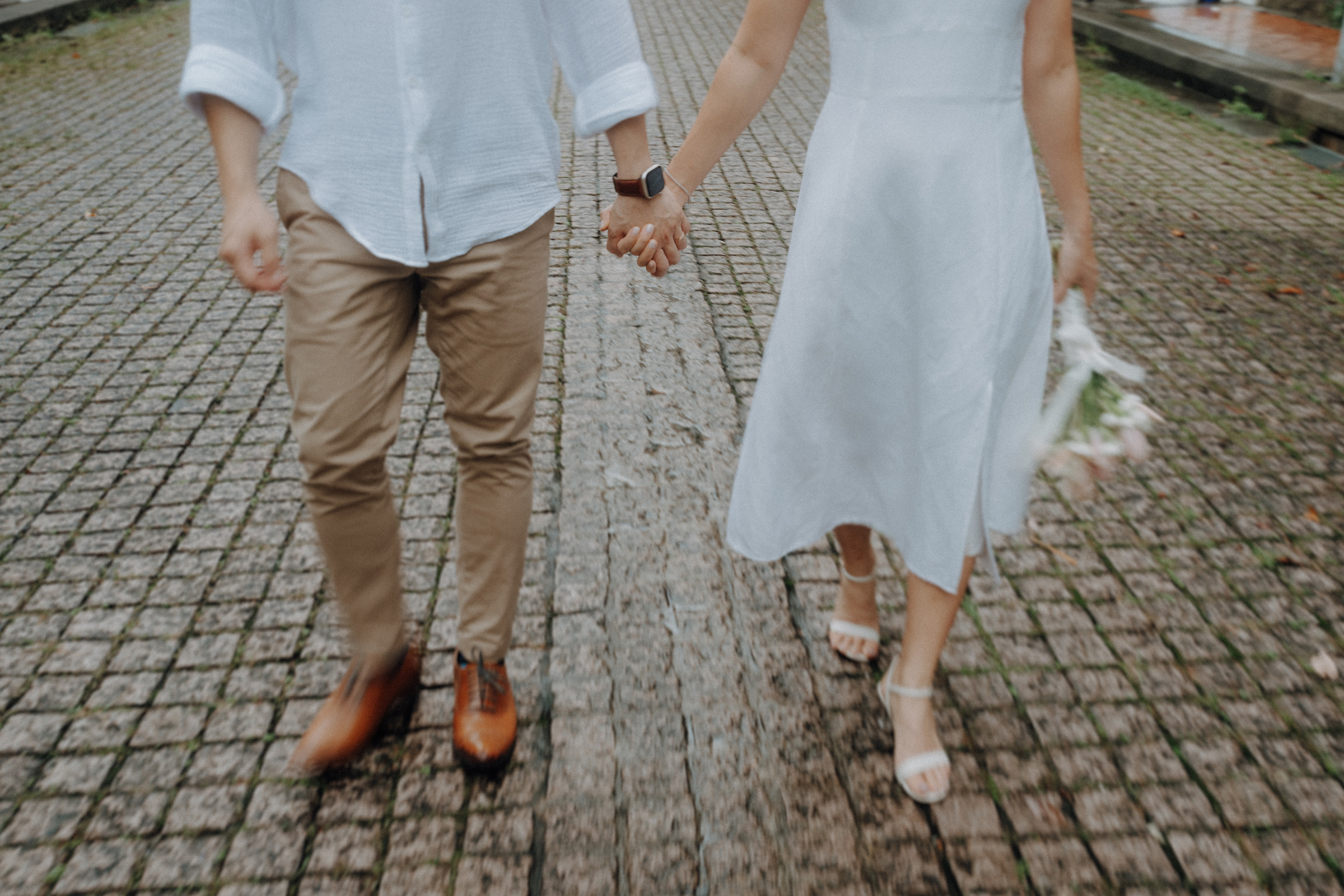 A couple walking hand in hand on a cobblestone path, with the woman holding a small bouquet.