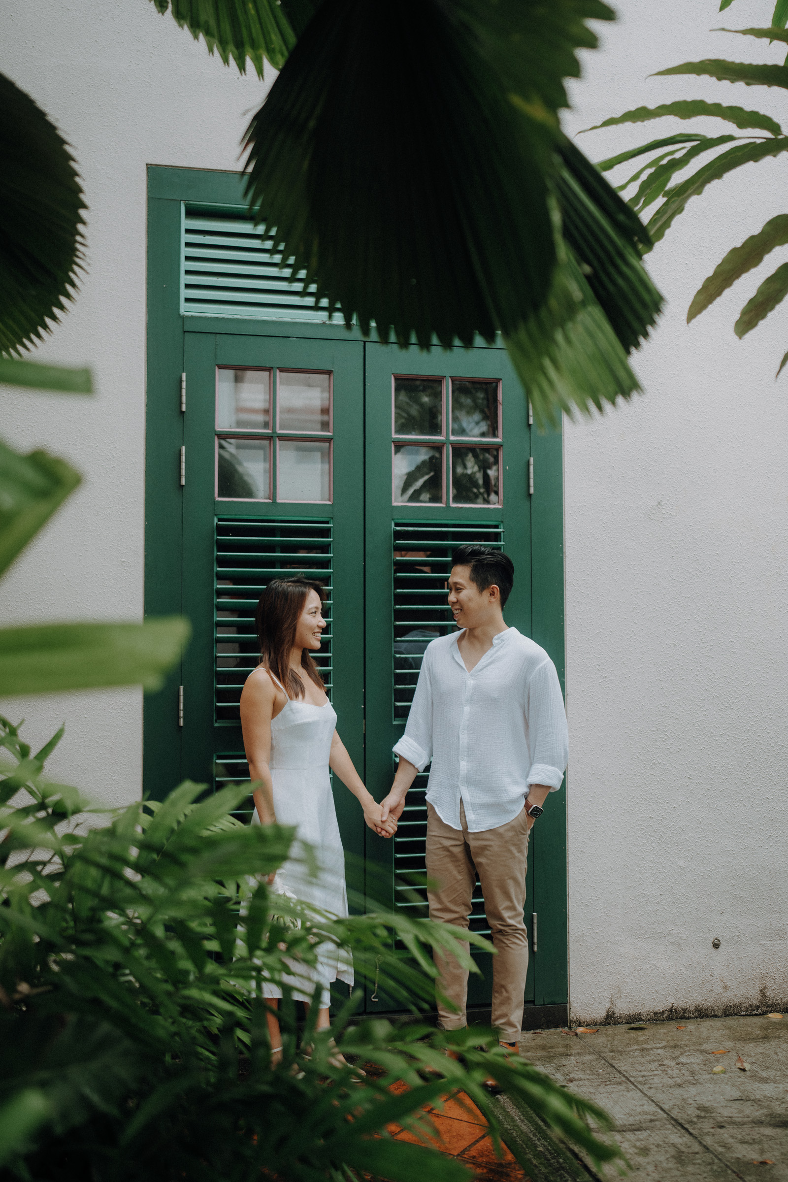 A couple holds hands while standing near a green shuttered window surrounded by lush greenery.
