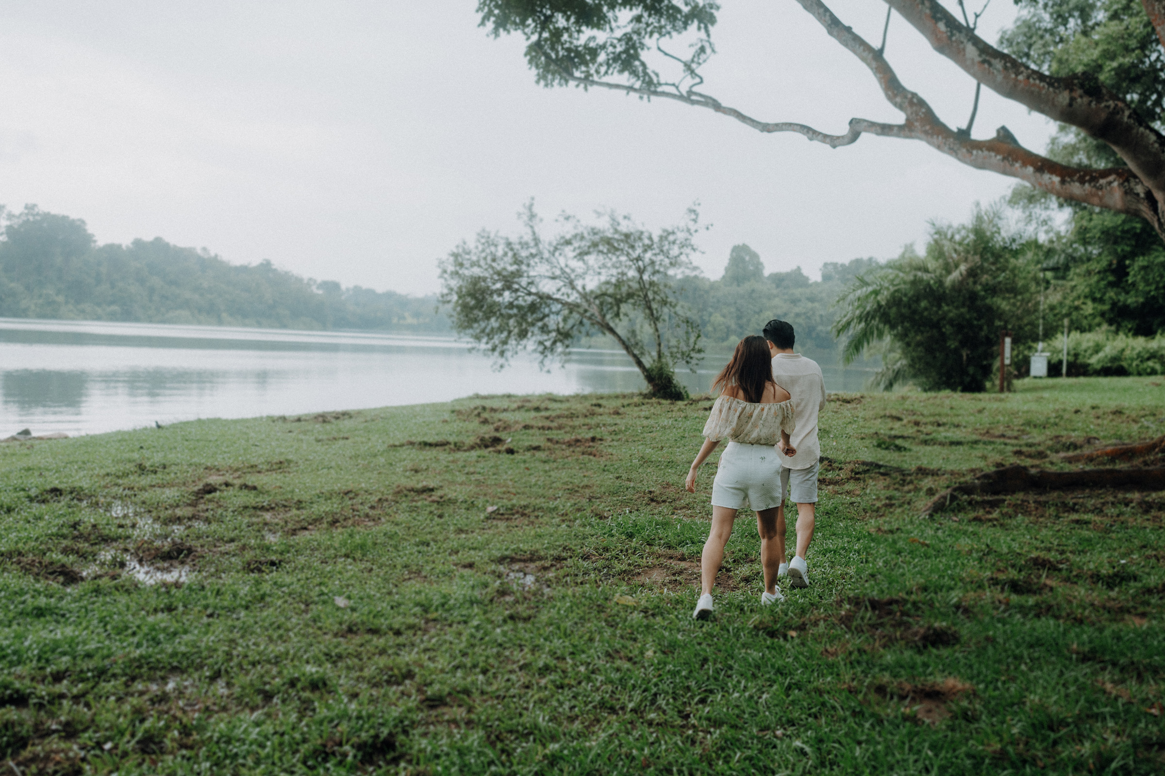 A couple walks hand in hand on a grassy lakeshore, surrounded by trees, with the lake visible to the left.
