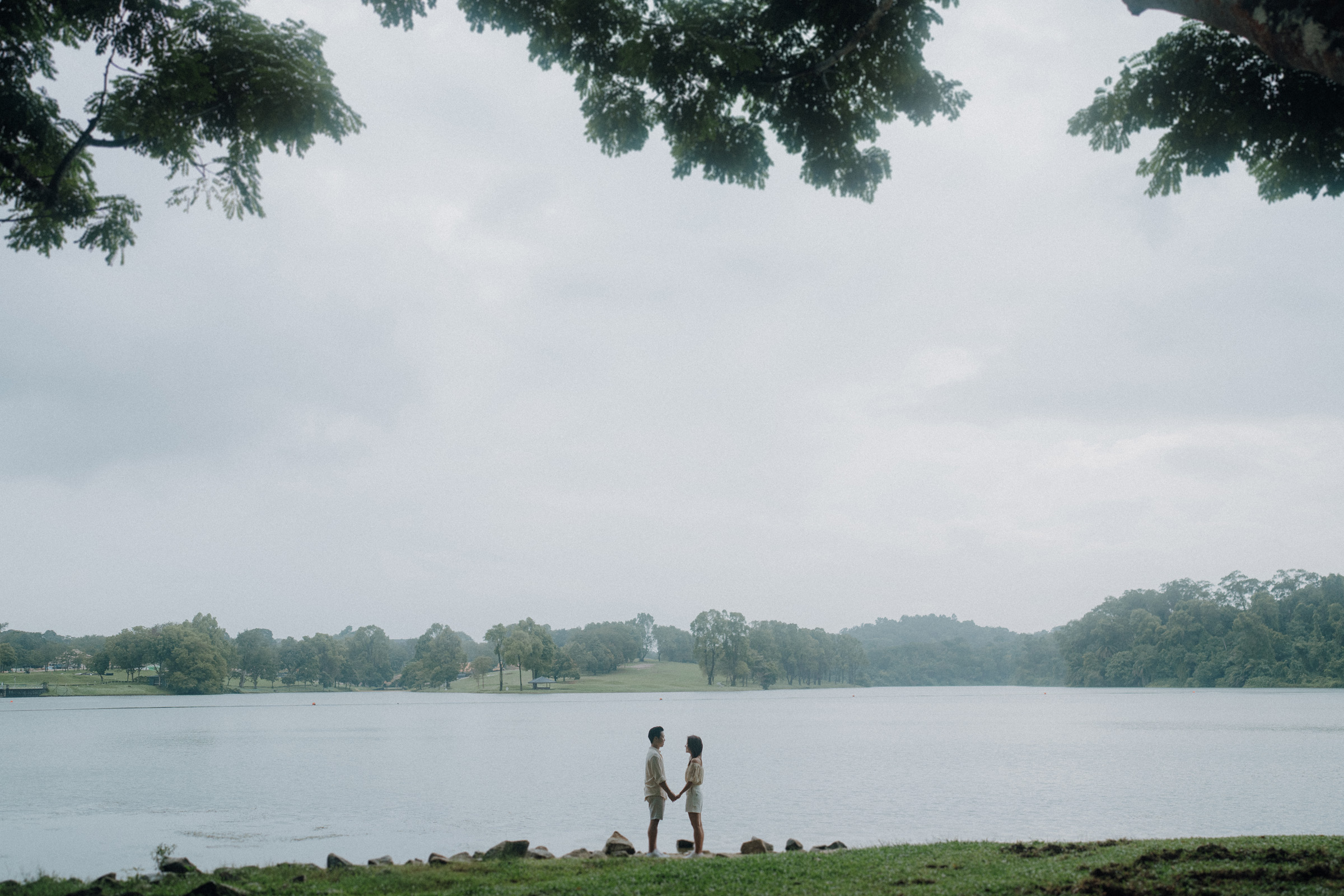 Two people stand by a lake holding hands, with trees in the foreground and a cloudy sky above.