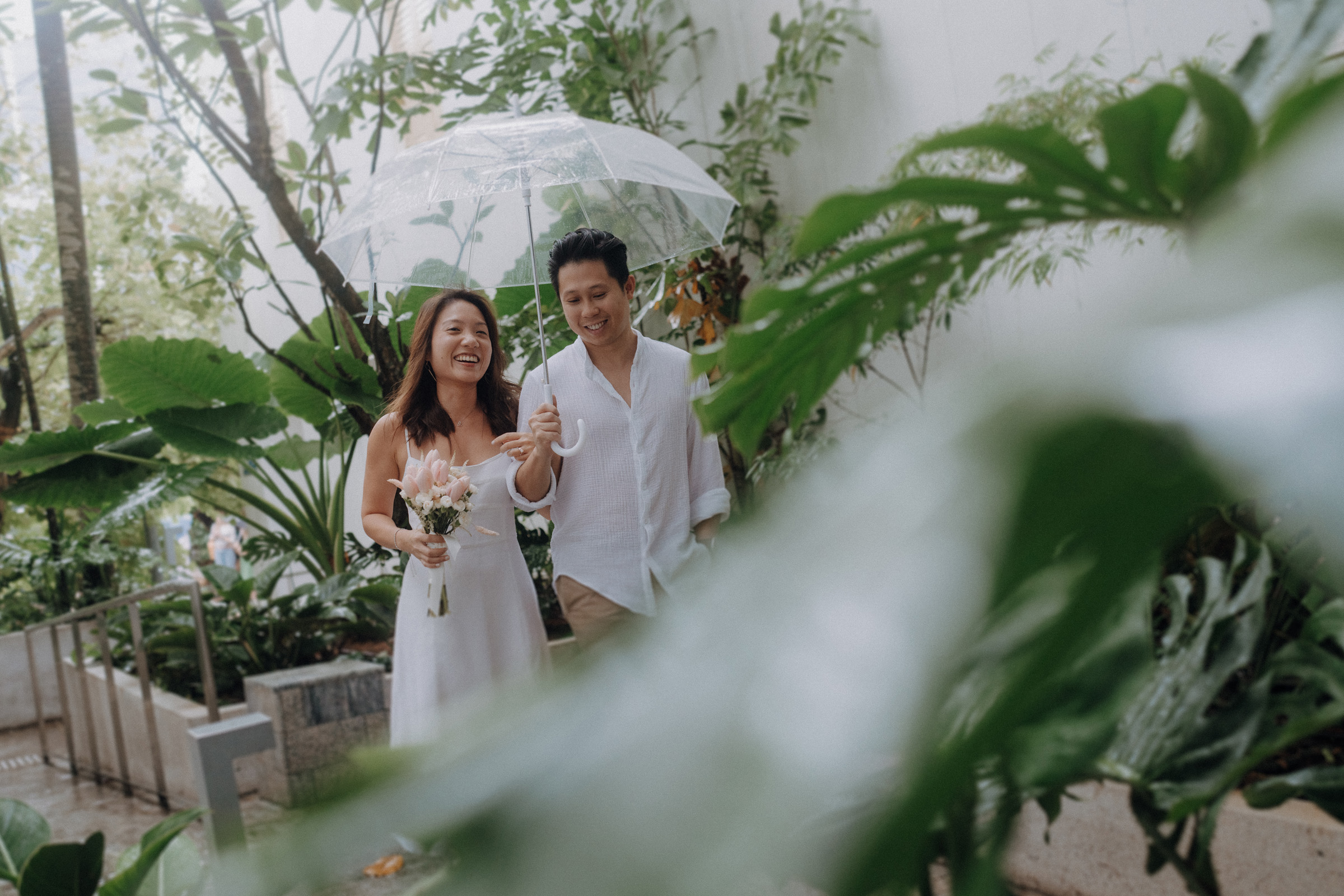 A couple walks under a clear umbrella, surrounded by lush green plants, with the woman holding a bouquet of flowers.