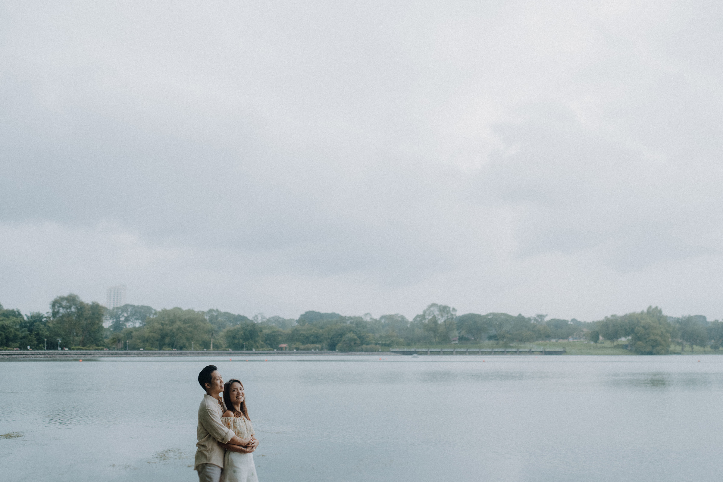 A couple stands by a calm lake under a cloudy sky, embracing each other. Trees and distant buildings are visible in the background.
