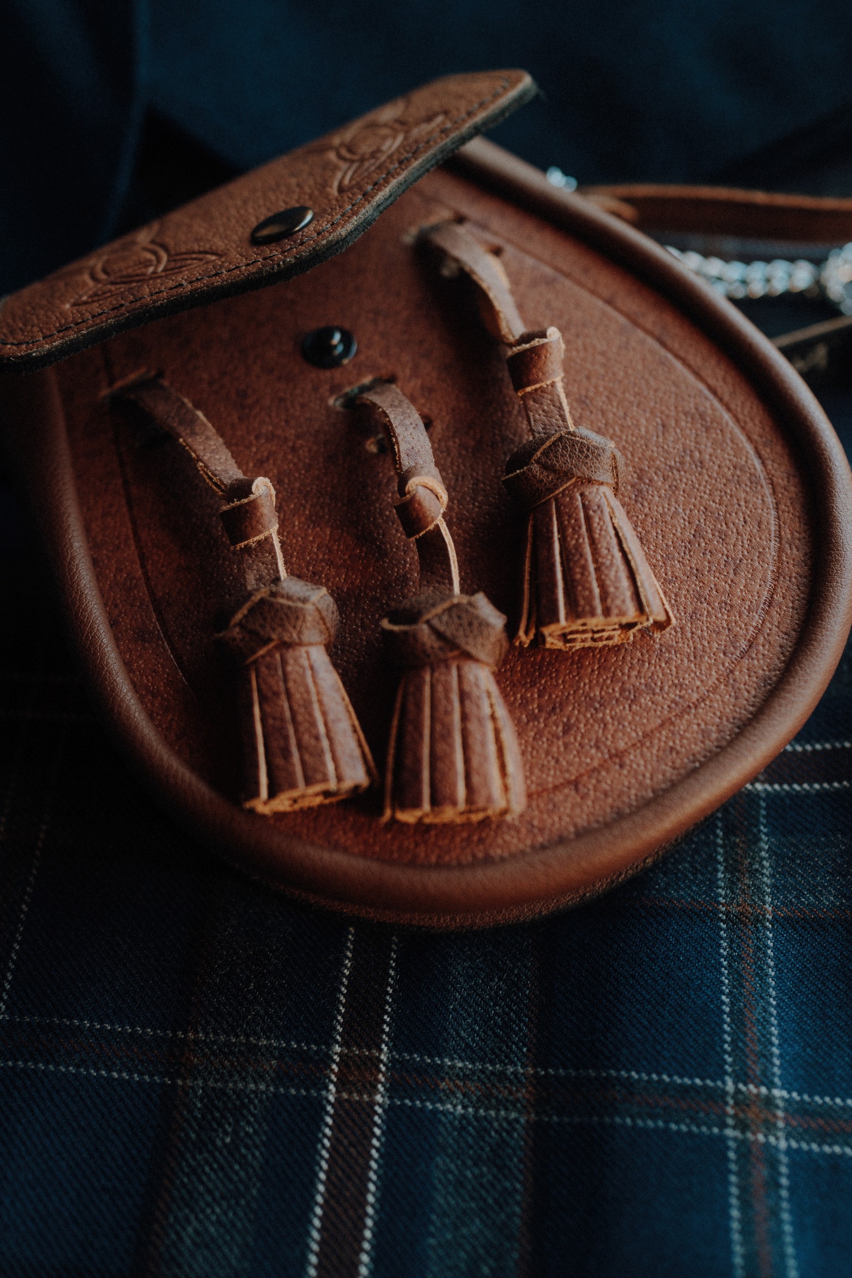 A brown leather sporran with three tassels rests on a dark plaid fabric background.