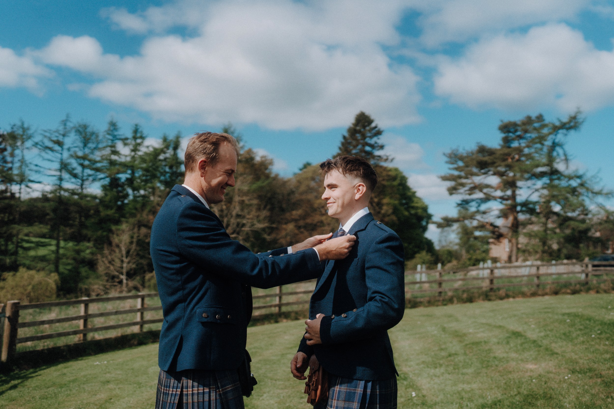 Two men wearing kilts and jackets stand outside on grass; one adjusts the other’s tie on a sunny day with trees and a fence in the background.