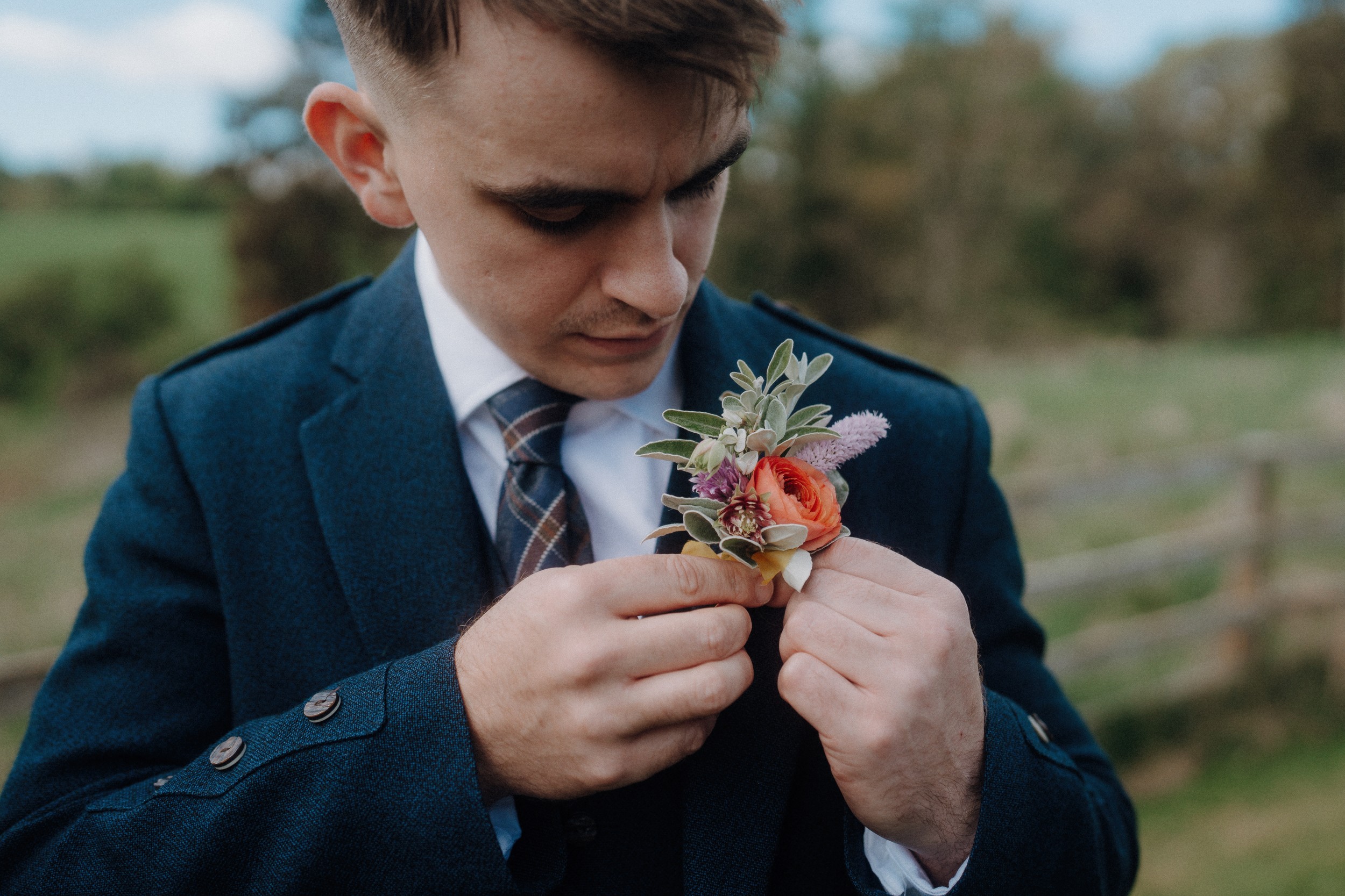 A man in a suit pins a colorful boutonniere with orange and purple flowers to his lapel outdoors.