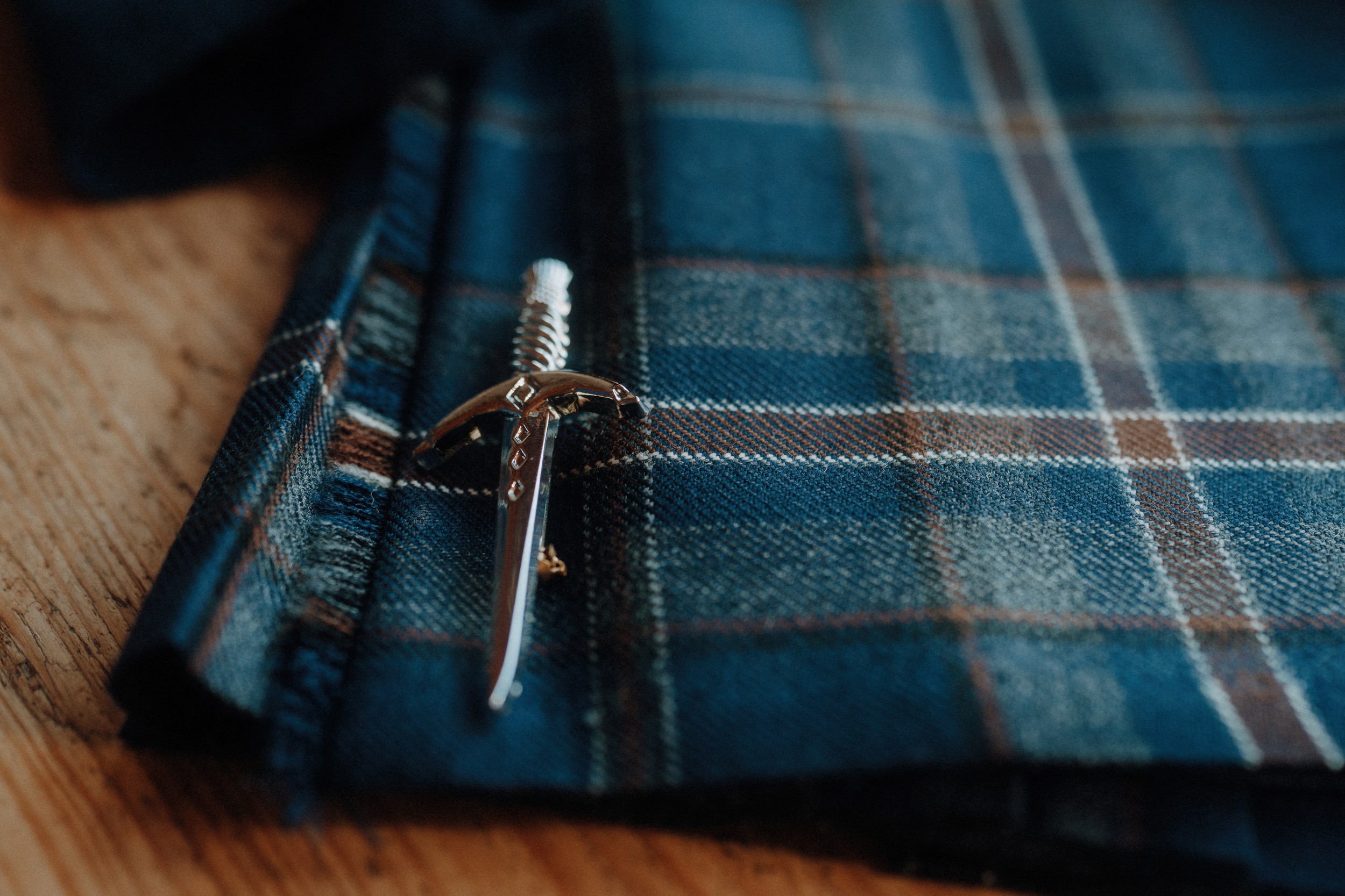 A silver dagger-shaped pin rests on a folded piece of blue plaid fabric on a wooden surface.