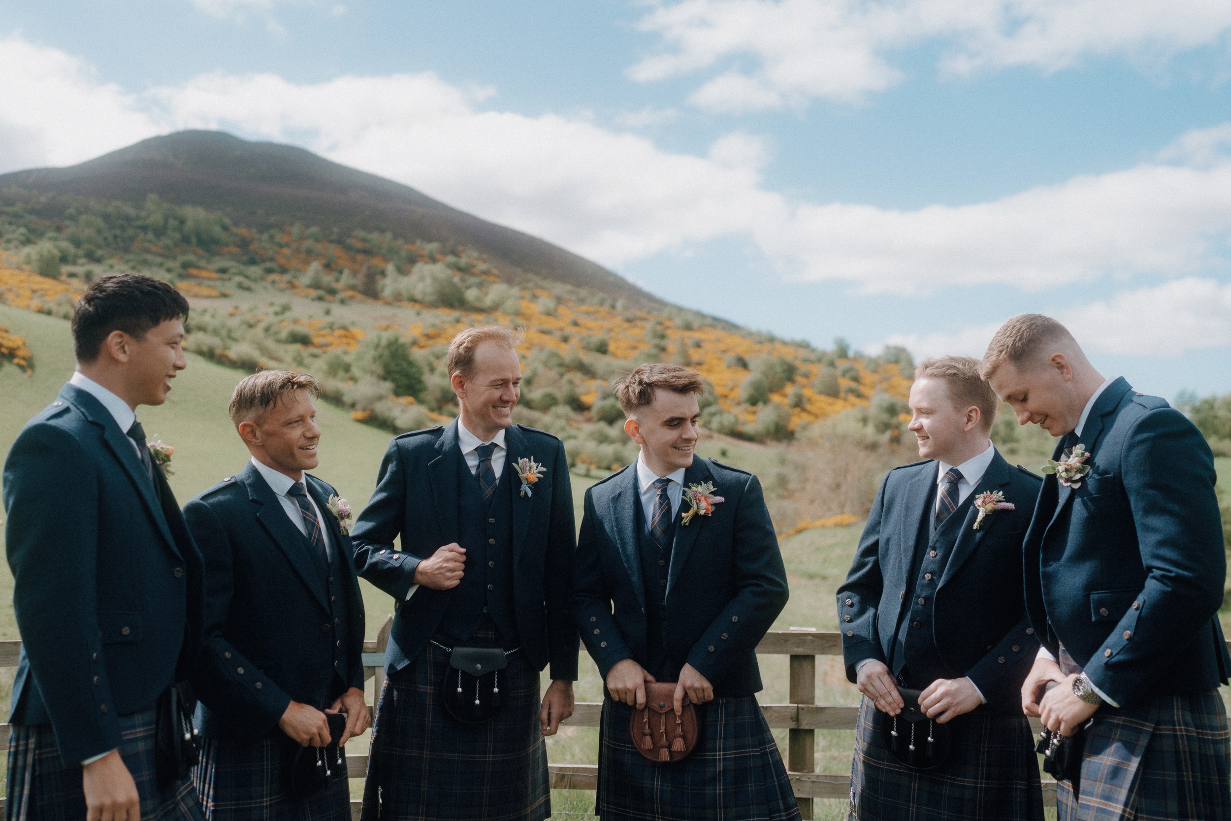 Six men in kilts and jackets stand outdoors by a wooden fence, with hills and greenery in the background under a partly cloudy sky.