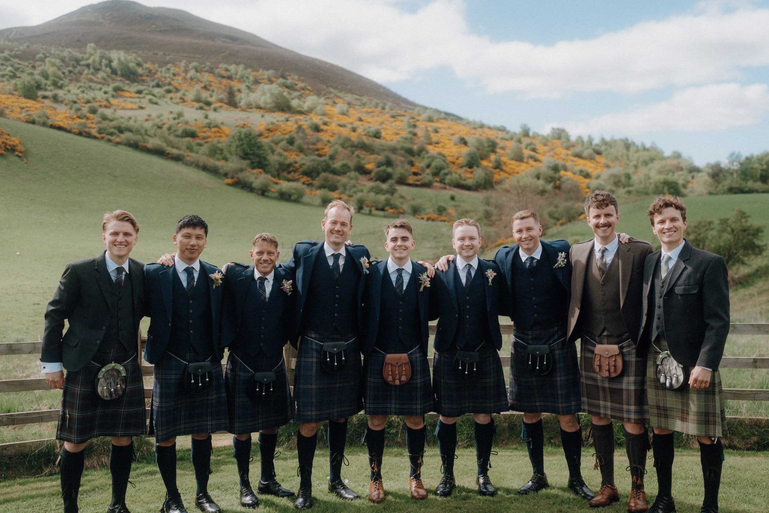 Nine men wearing kilts and jackets stand side by side outdoors in front of a wooden fence, with green hills and yellow flowers in the background.