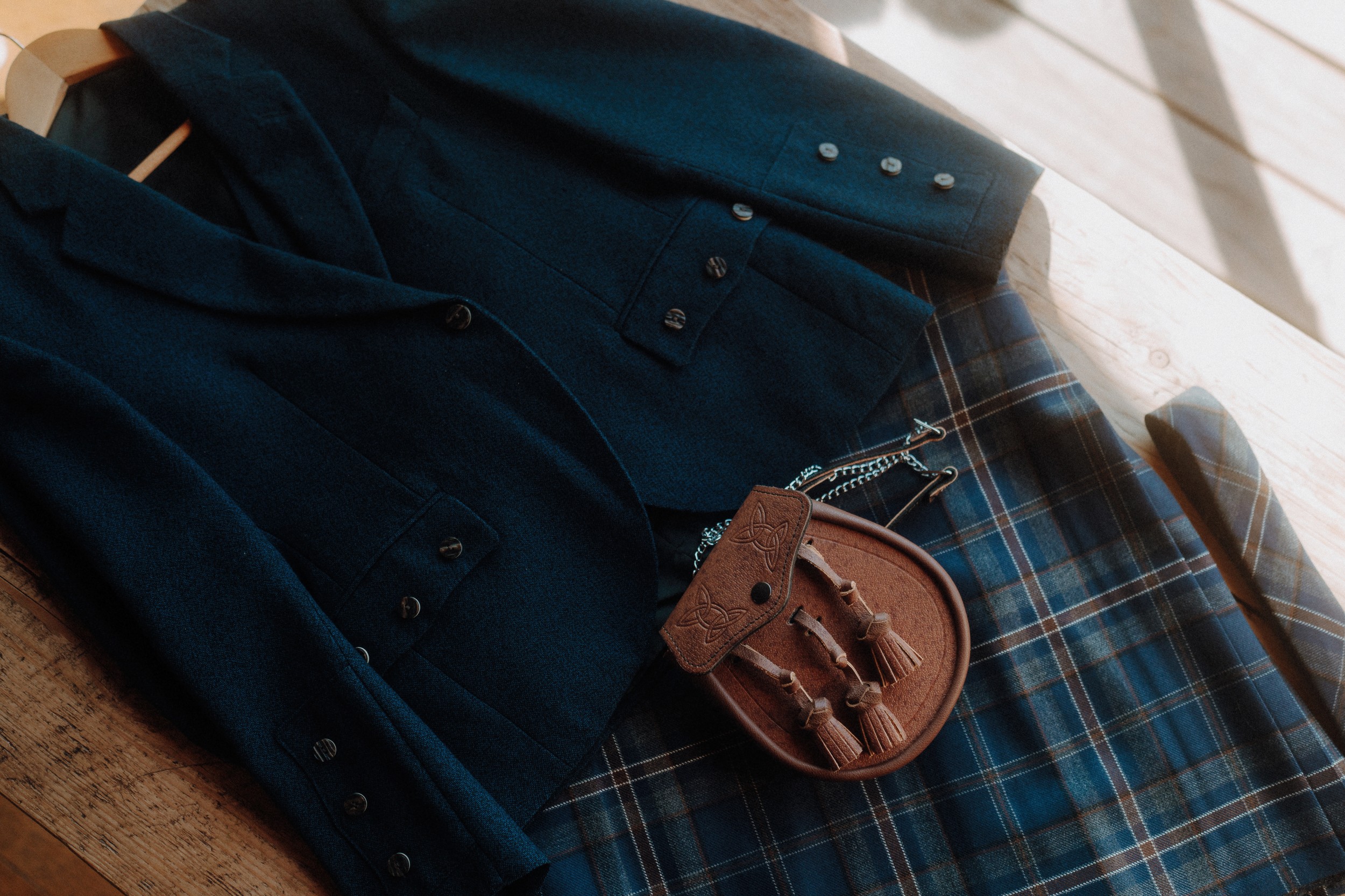 A dark blue blazer and a blue tartan plaid kilt are laid out on a wooden surface, accompanied by a brown sporran with tassels.