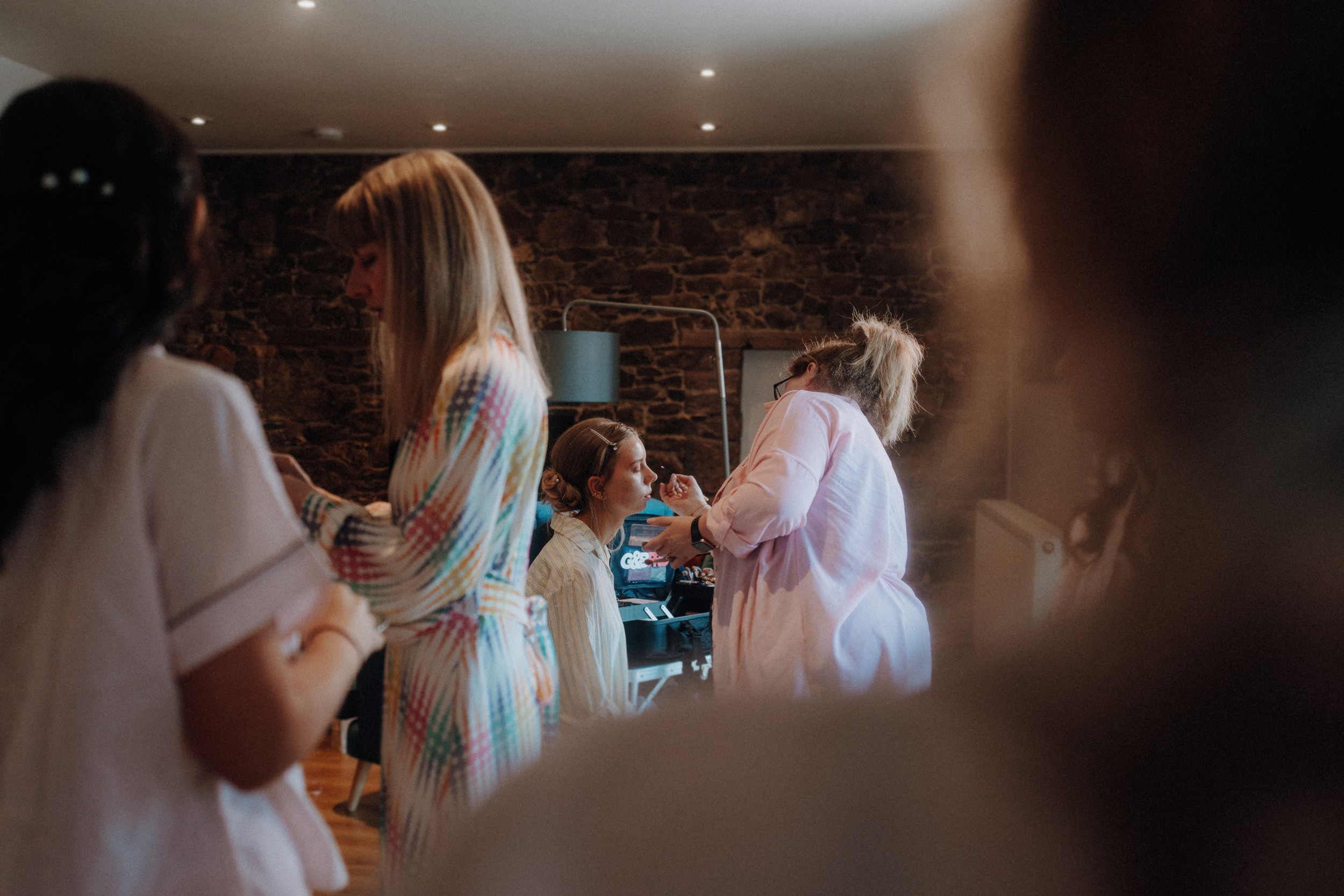 A group of women in a softly lit room, with one woman seated while another applies her makeup. Others are standing nearby, partially out of focus.
