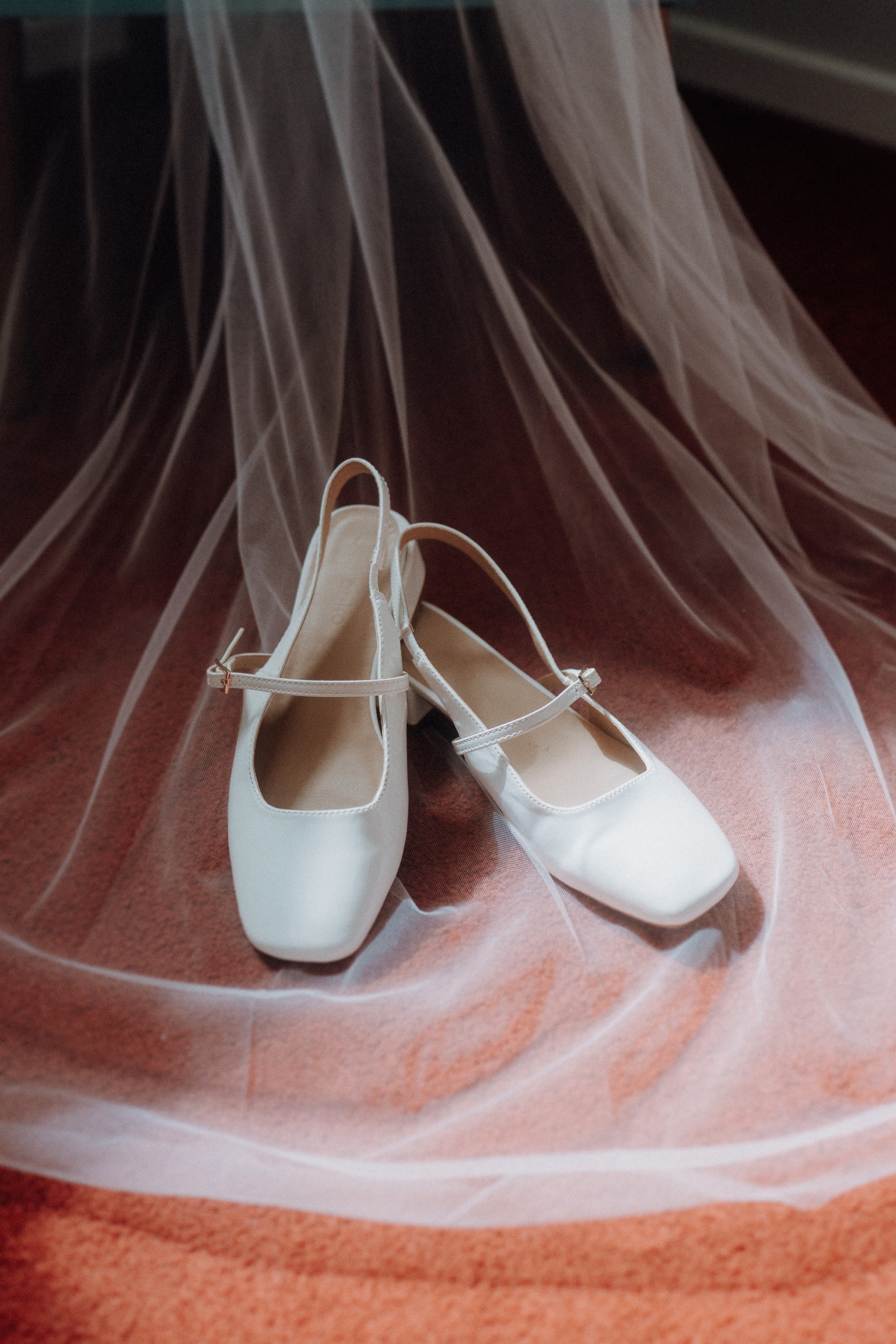 A pair of white, closed-toe, slingback shoes with straps sit on a pink carpet, partially covered by sheer white fabric.
