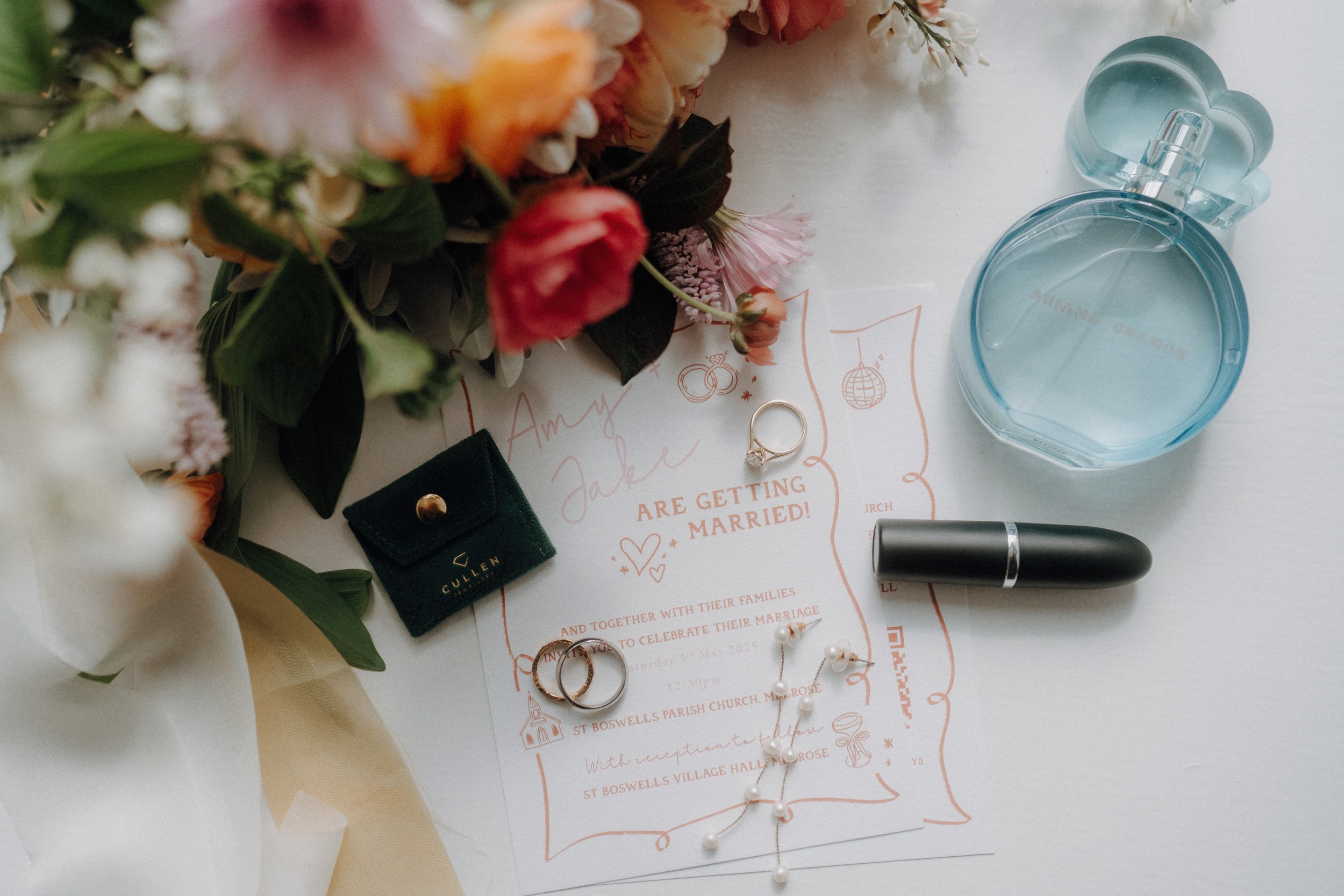 Flat lay of wedding invitation with gold rings, a necklace, black lipstick, perfume bottle, ring box, and a bouquet of flowers arranged on a white surface.