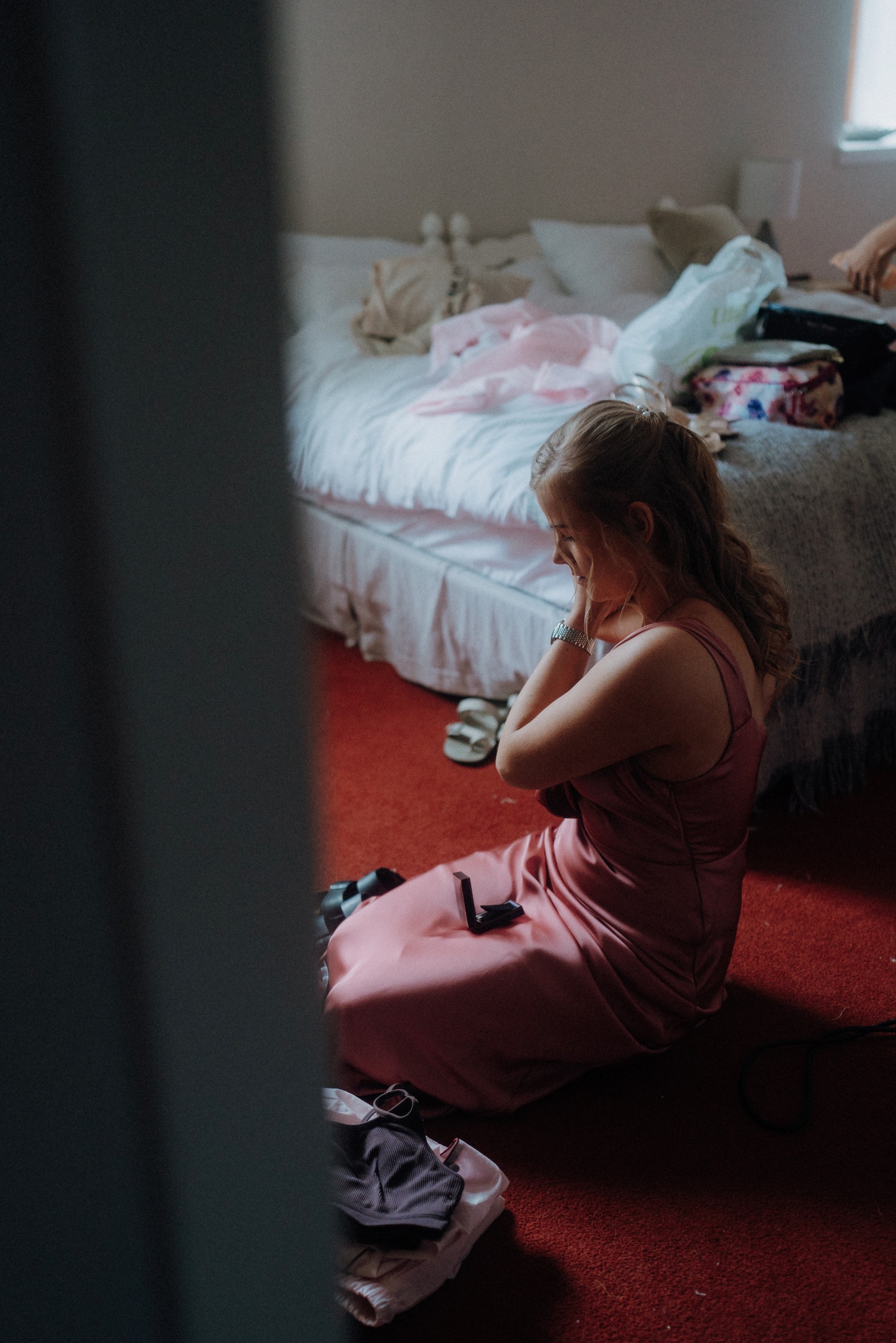 A woman in a pink dress sits on the floor adjusting jewelry in a bedroom with a red carpet and a bed in the background.