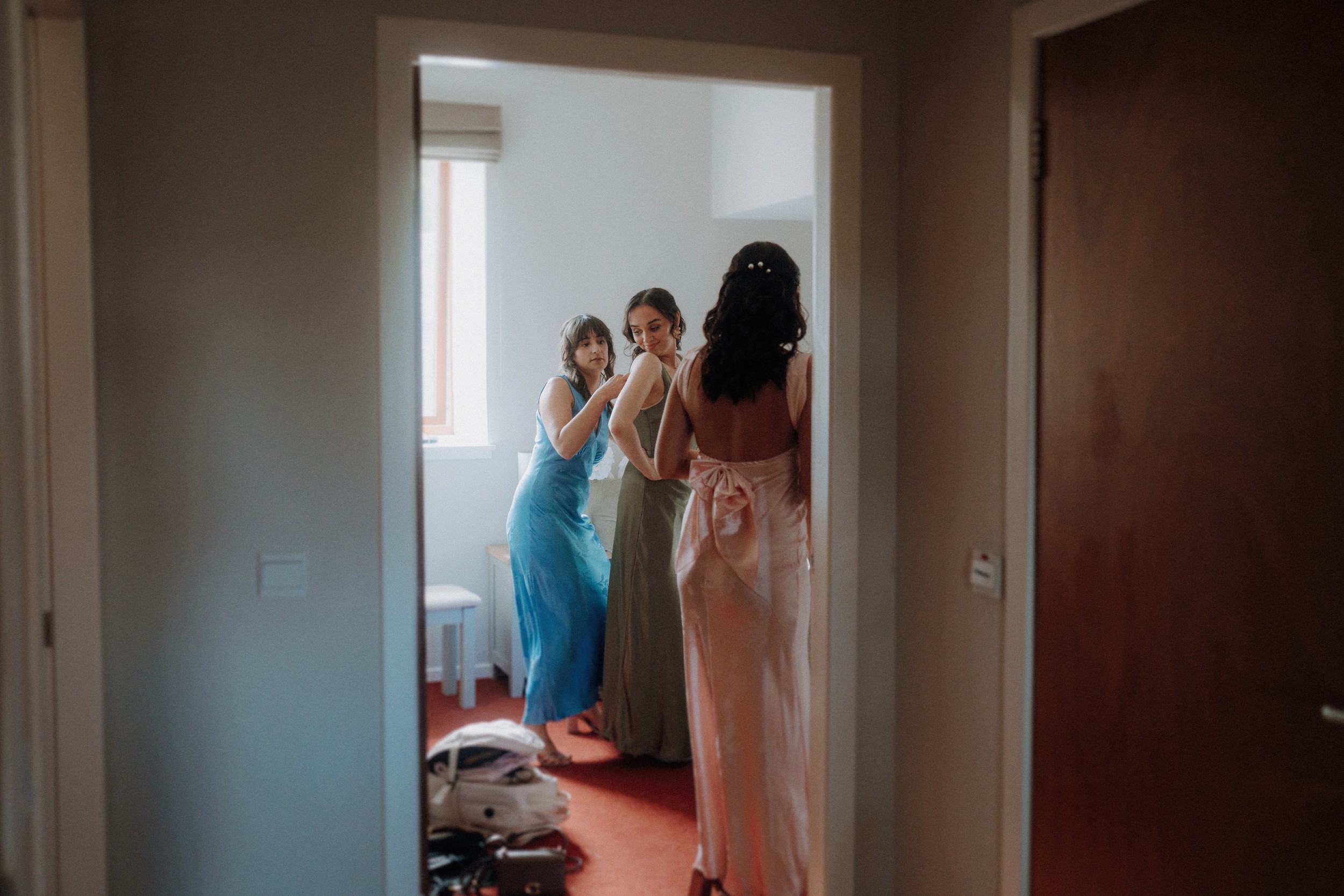 Three women in formal dresses stand in a room, two are looking in a mirror while the third stands with her back to the camera. A bag sits on the floor near them.