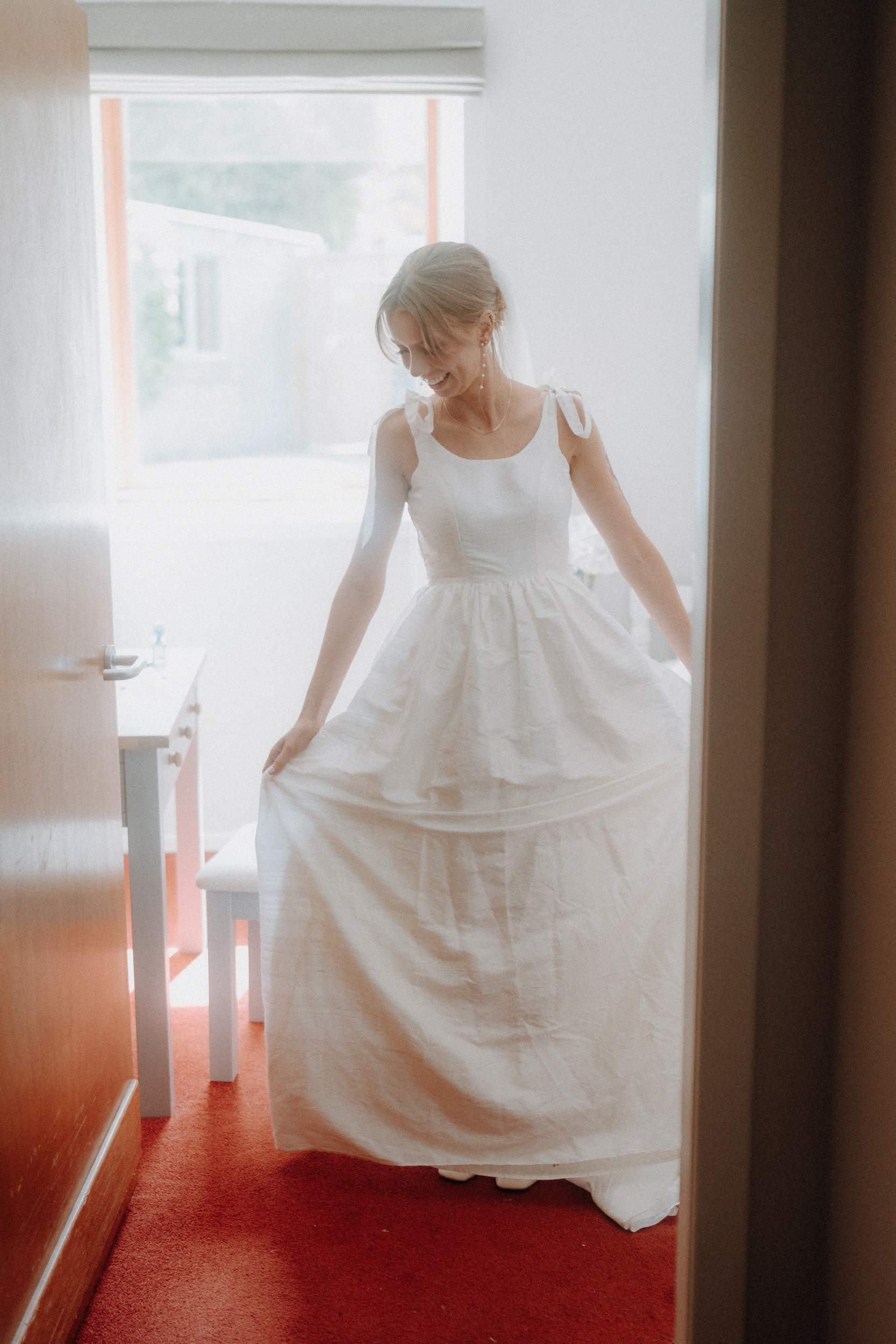 Woman in a sleeveless white dress stands indoors on a red carpet, holding the sides of her skirt and looking down, near a table and window with natural light.