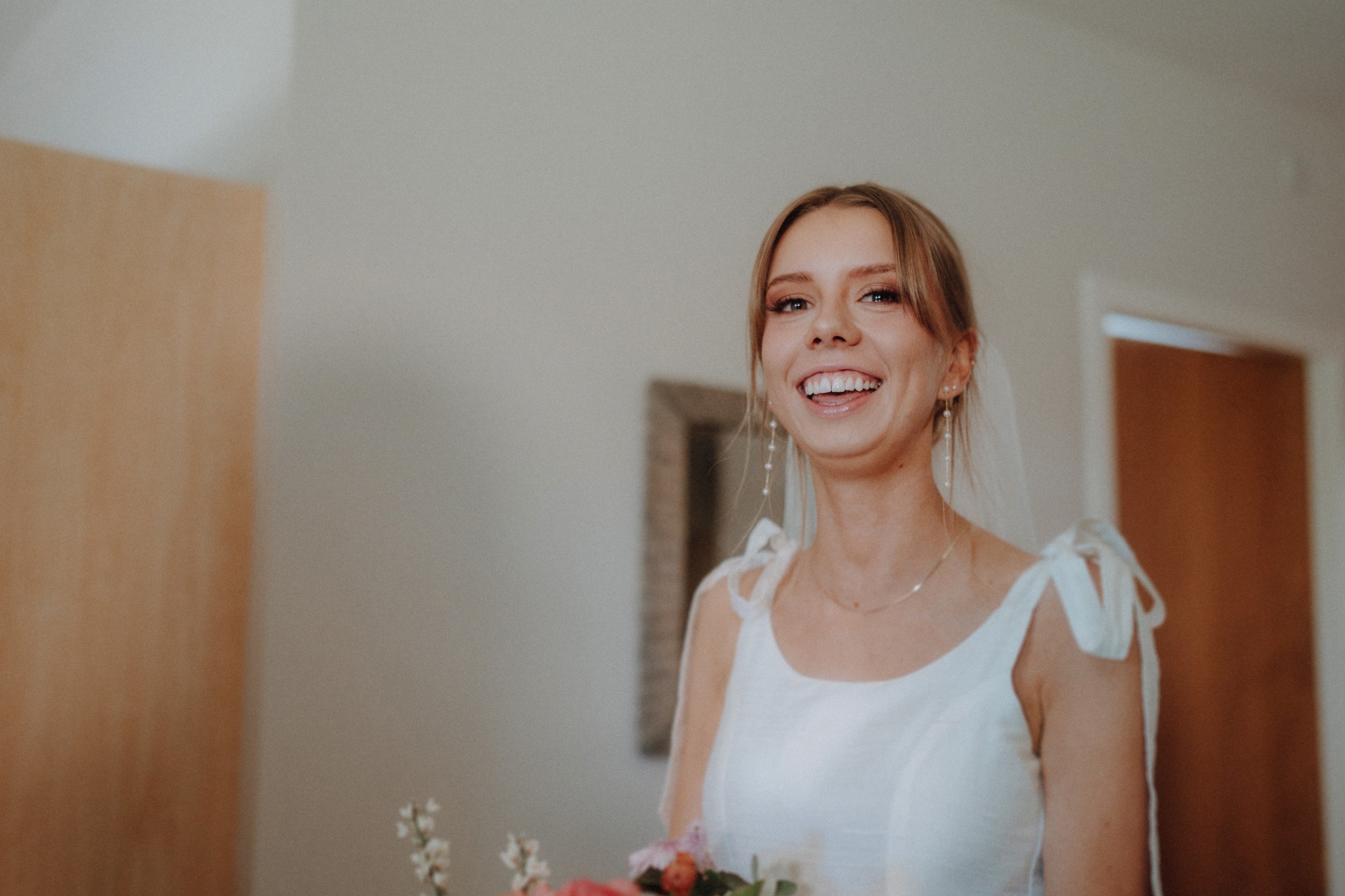 A woman in a white dress smiles while holding a bouquet of flowers indoors, standing near wooden doors and a light-colored wall.